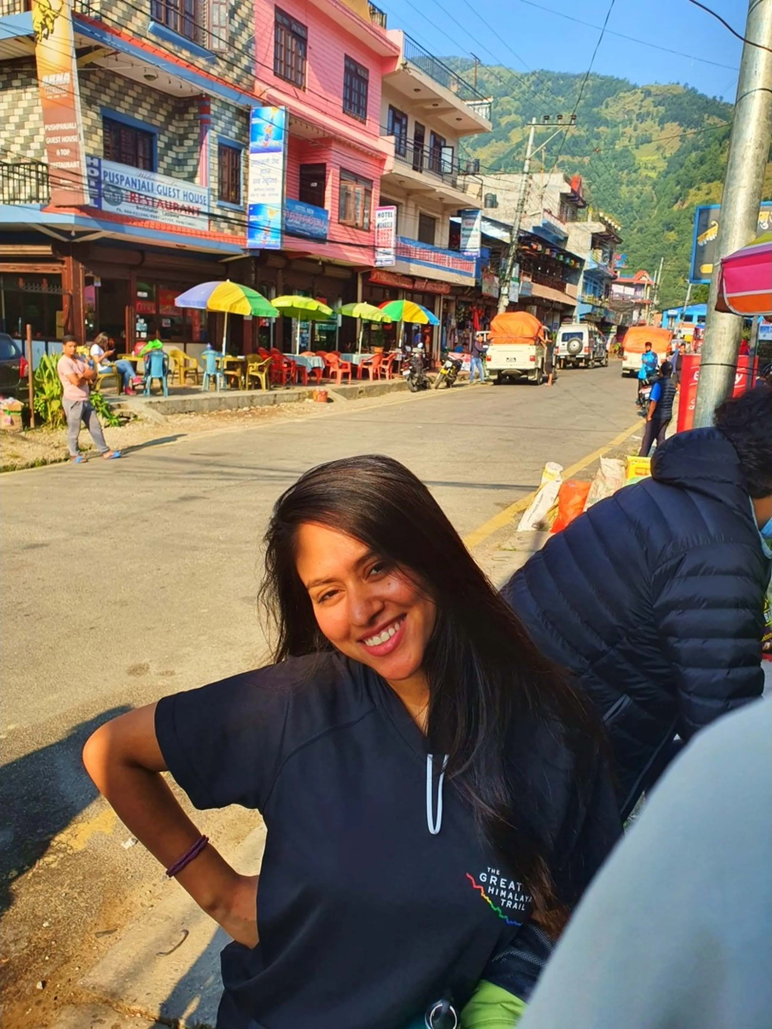 A young Nepali woman in a navy blue Great Himalayan Trail branded t-shirt smiles radiantly at the camera with hands on hips, standing on a busy mountain town street in Nepal - behind her a row of colorful buildings including a pink and blue guest house with a 'Puspanjali Guest House' sign, outdoor restaurants with yellow and green umbrellas, motorcycles, a truck, and other travelers - green forested hills rising in the background under morning sunlight - a vibrant and energetic Nepal trekking town street scene capturing the buzz of departure day on the Annapurna Circuit