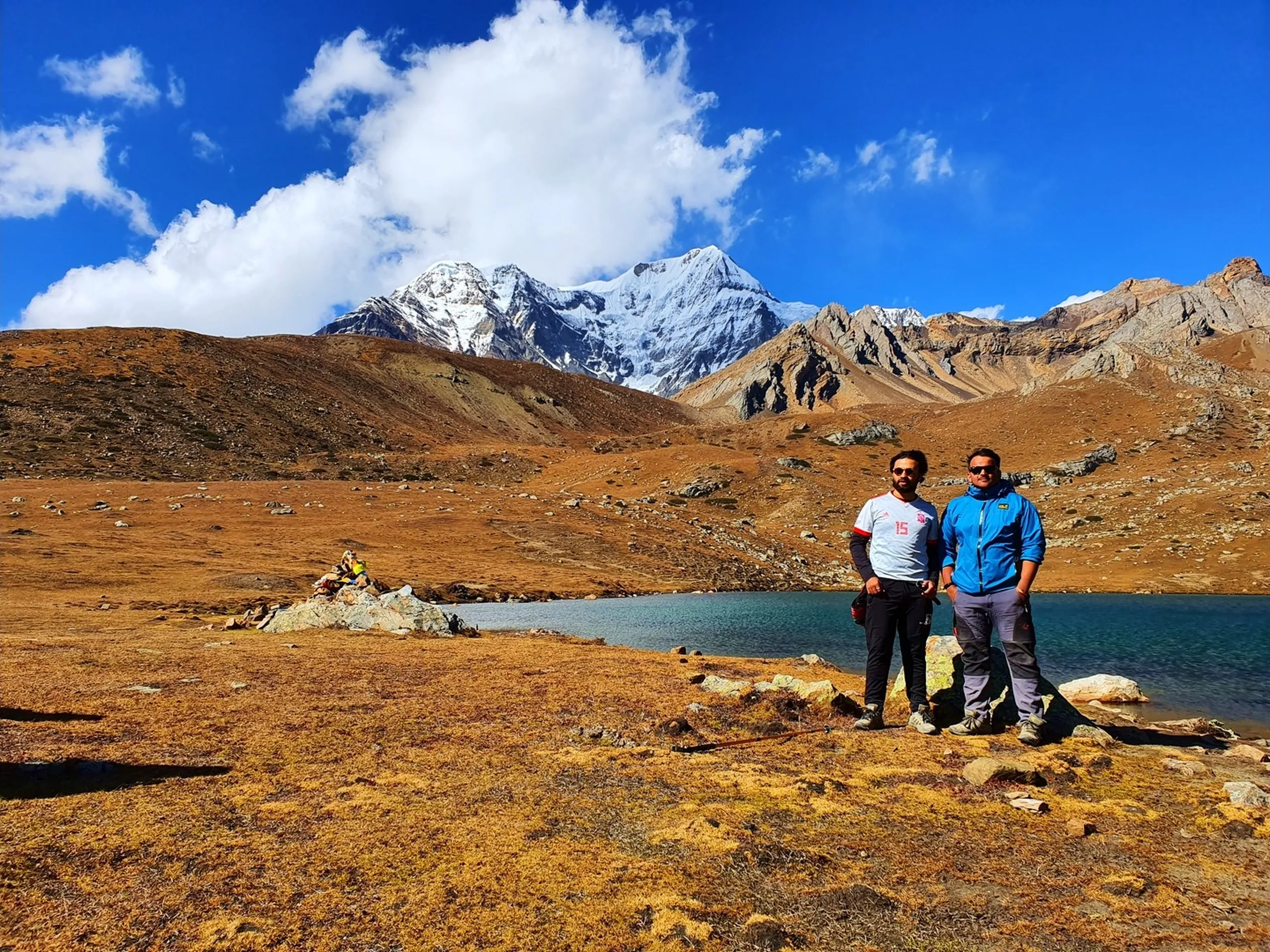 Two male trekkers standing together on the golden alpine grassland at Ice Lake Nepal at 4600 metres, one in a white football jersey with red scarf and the other in a blue Jack Wolfskin jacket and sunglasses, a stone prayer flag cairn to their left, the vivid blue-green glacial lake behind them, and a striking snow-capped Himalayan peak rising above rust-brown ridges under a blue sky with dramatic white clouds - Ice Lake Nepal trekking duo, Annapurna Circuit Ice Lake friends, Nepal high altitude lake 4600m