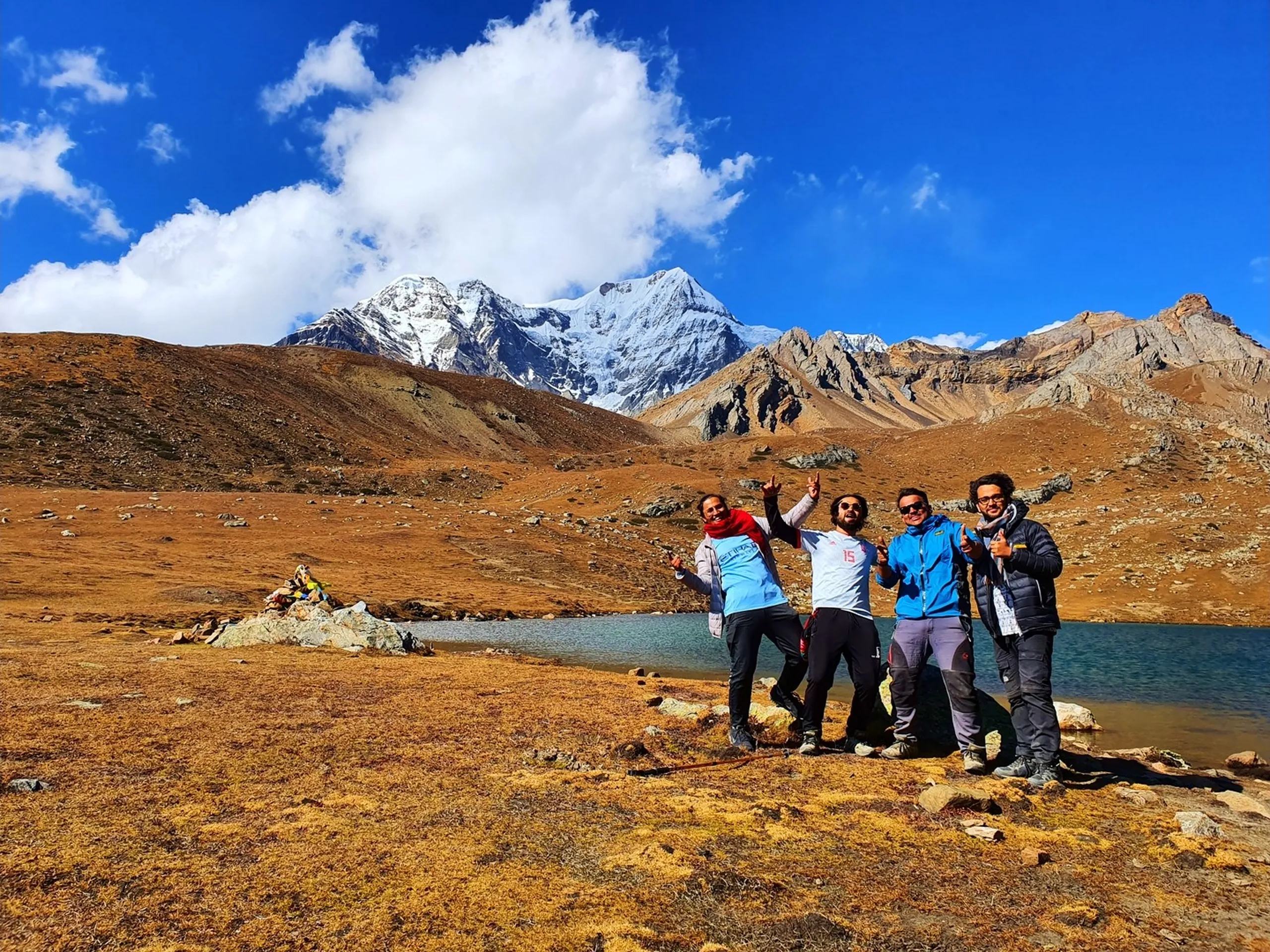 Four friends posing joyfully at Ice Lake Manang Nepal at high altitude alpine lake trekking destination on Annapurna Circuit Trek with snow-capped Himalayan peaks Gangapurna and Annapurna III towering in background under vivid cobalt blue sky with dramatic white clouds golden autumn tundra meadows surrounding glacial turquoise lake Buddhist prayer flags stone cairn visible trekkers celebrating milestone achievement Nepal mountain adventure