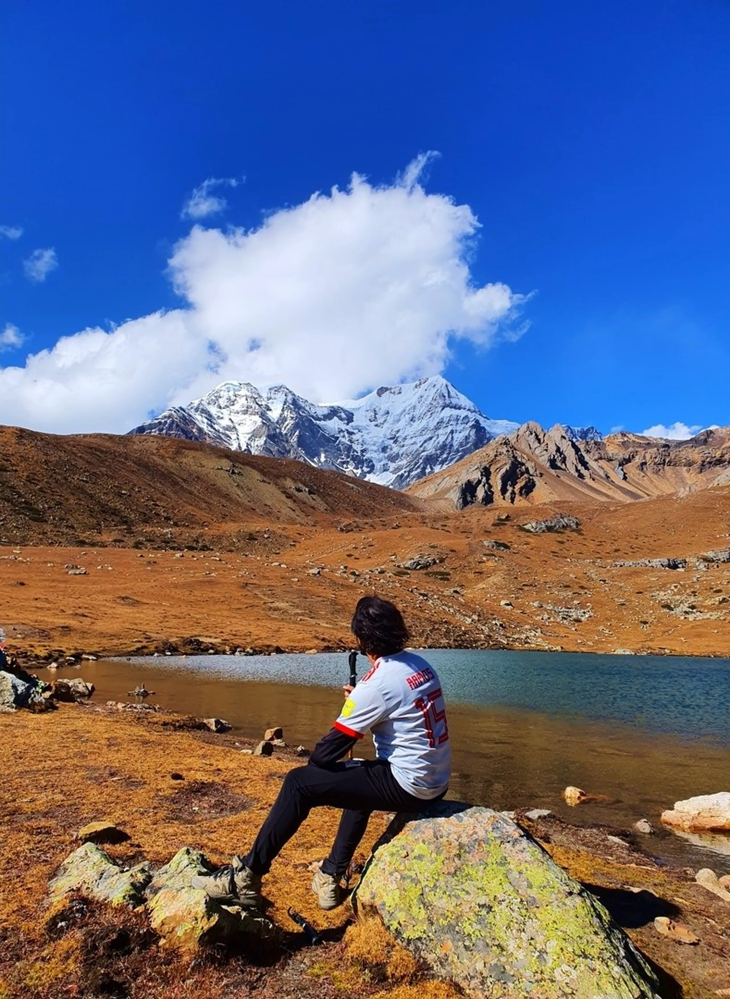 Solo male trekker sitting on lichen-covered boulder at Ice Lake Manang Nepal contemplating breathtaking panoramic view of snow-covered Himalayan mountain peaks Gangapurna glacier reflected in glacial turquoise alpine lake golden tundra terrain autumn colors on Annapurna Circuit Trek wearing white sports jersey number 15 with trekking poles Nepal high altitude adventure travel photography