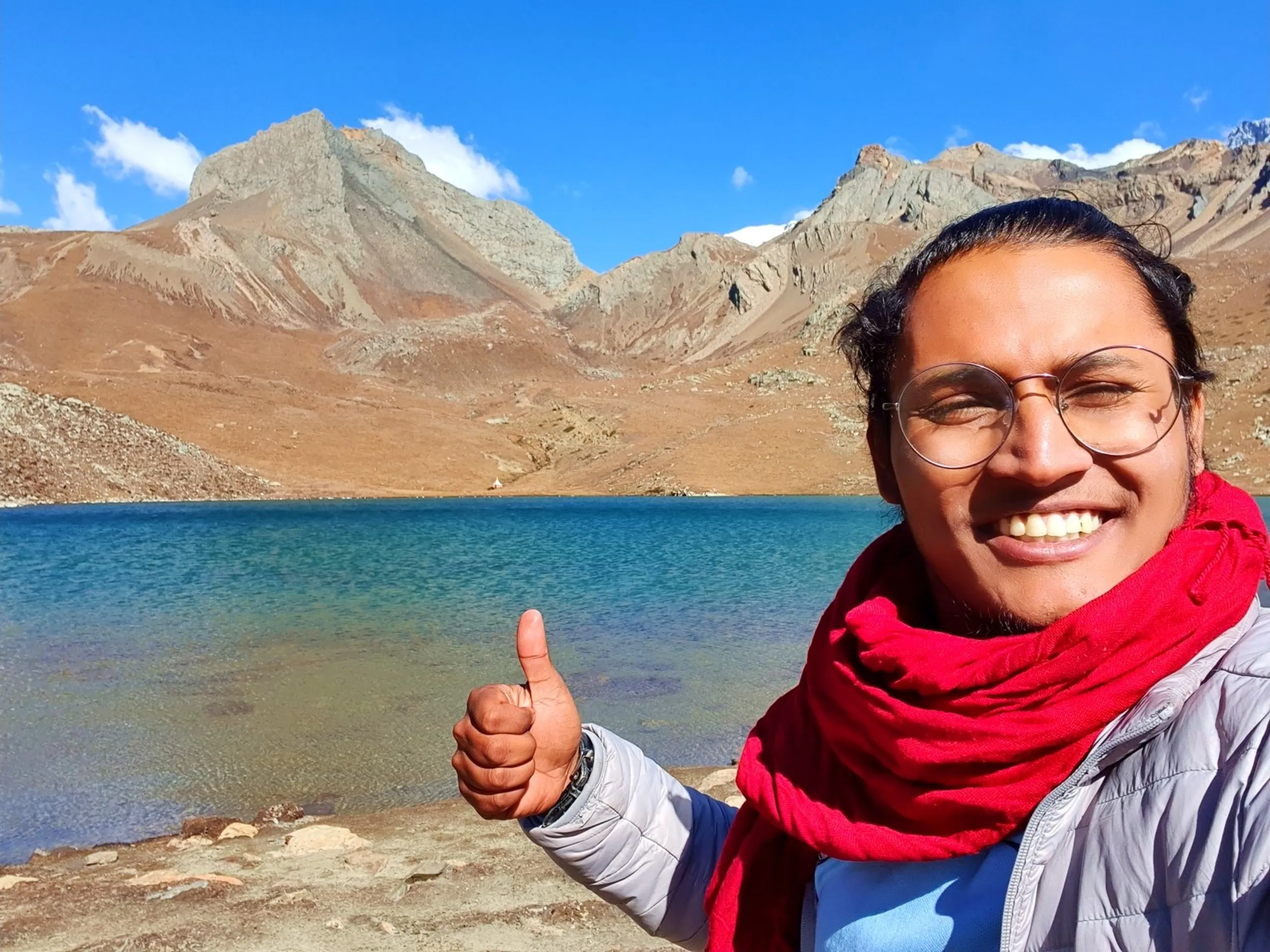 Close-up selfie of smiling male trekker with round glasses and red scarf giving thumbs up at Ice Lake Manang Nepal with crystal clear turquoise glacial lake and dramatic rocky grey Himalayan mountain ridgeline in background bright blue sky Annapurna Circuit Trek achievement Nepal adventure travel happiness high altitude trekking success