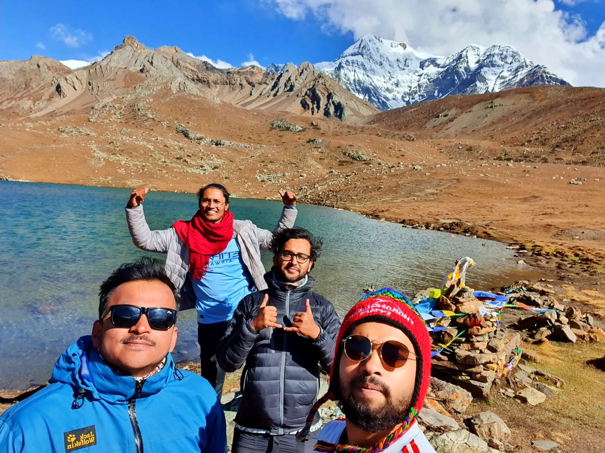 Group selfie of four trekking friends at Ice Lake Manang Nepal 4600m altitude Annapurna Circuit with massive snow-capped Himalayan peaks Gangapurna in background one trekker wearing Jack Wolfskin blue jacket sunglasses mustache another in red scarf grey jacket raising arms in celebration third in grey puffer jacket giving thumbs up fourth in colorful red knit Nepali thokar hat sunglasses with vibrant Buddhist prayer flags and stone cairn beside glacial lake Nepal mountain trekking group travel