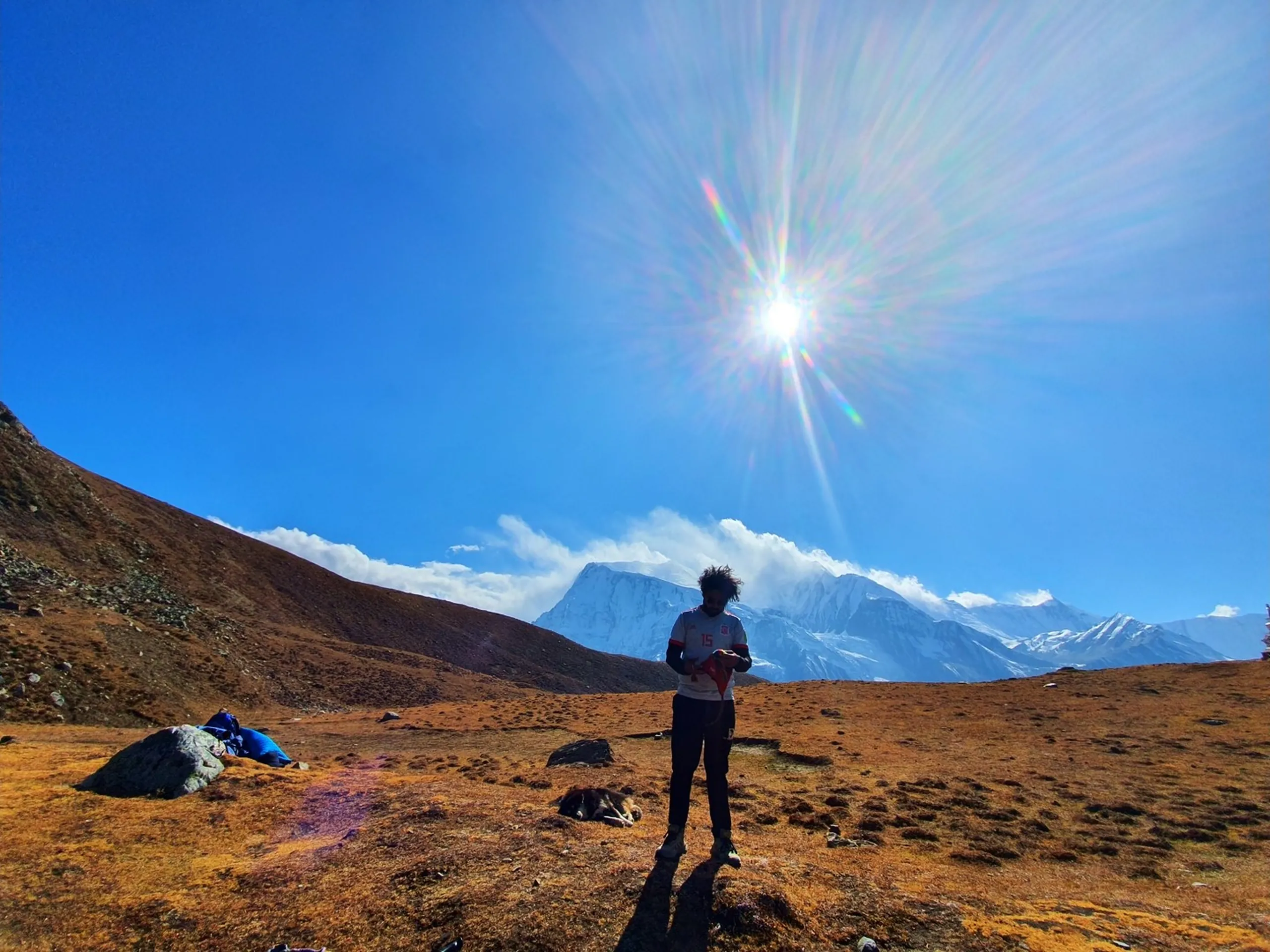 Lone trekker standing silhouetted against brilliant sunburst lens flare in deep blue sky on golden orange high altitude alpine meadow Manang Nepal Annapurna region with dramatic snow-covered Himalayan mountain range and billowing clouds in background wearing white sports jersey number 15 trekking gear bags visible on ground Nepal high altitude trek preparation Annapurna Circuit adventure travel