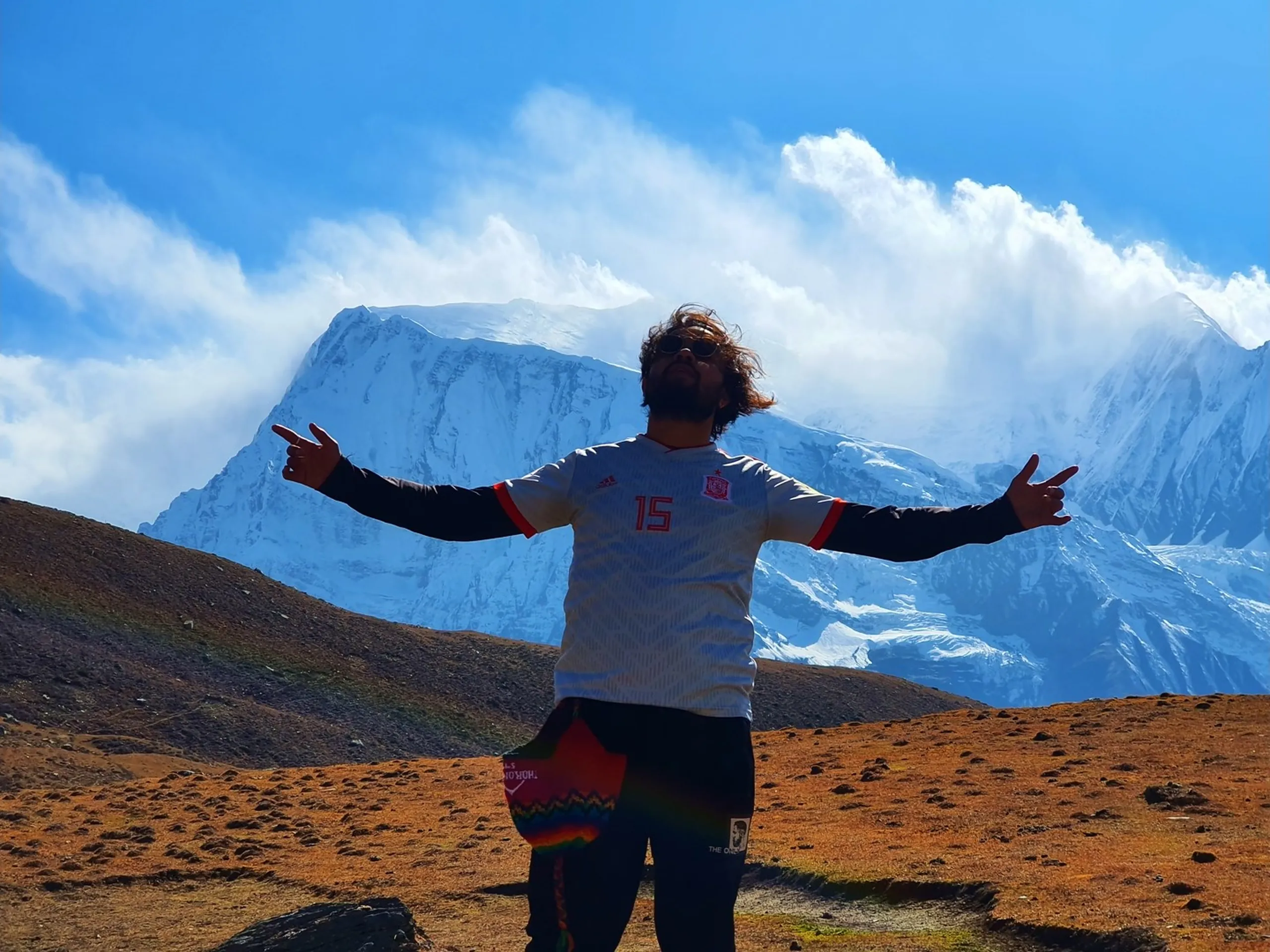 Male trekker with long curly hair and sunglasses wearing white Adidas Spain football jersey number 15 standing arms outstretched wide in triumphant victory pose against massive close-up snow-covered Himalayan mountain peaks Gangapurna glacier dramatic cloud formations Manang Nepal Annapurna Circuit Trek euphoria freedom mountains high altitude adventure travel milestone Nepal trekking blog