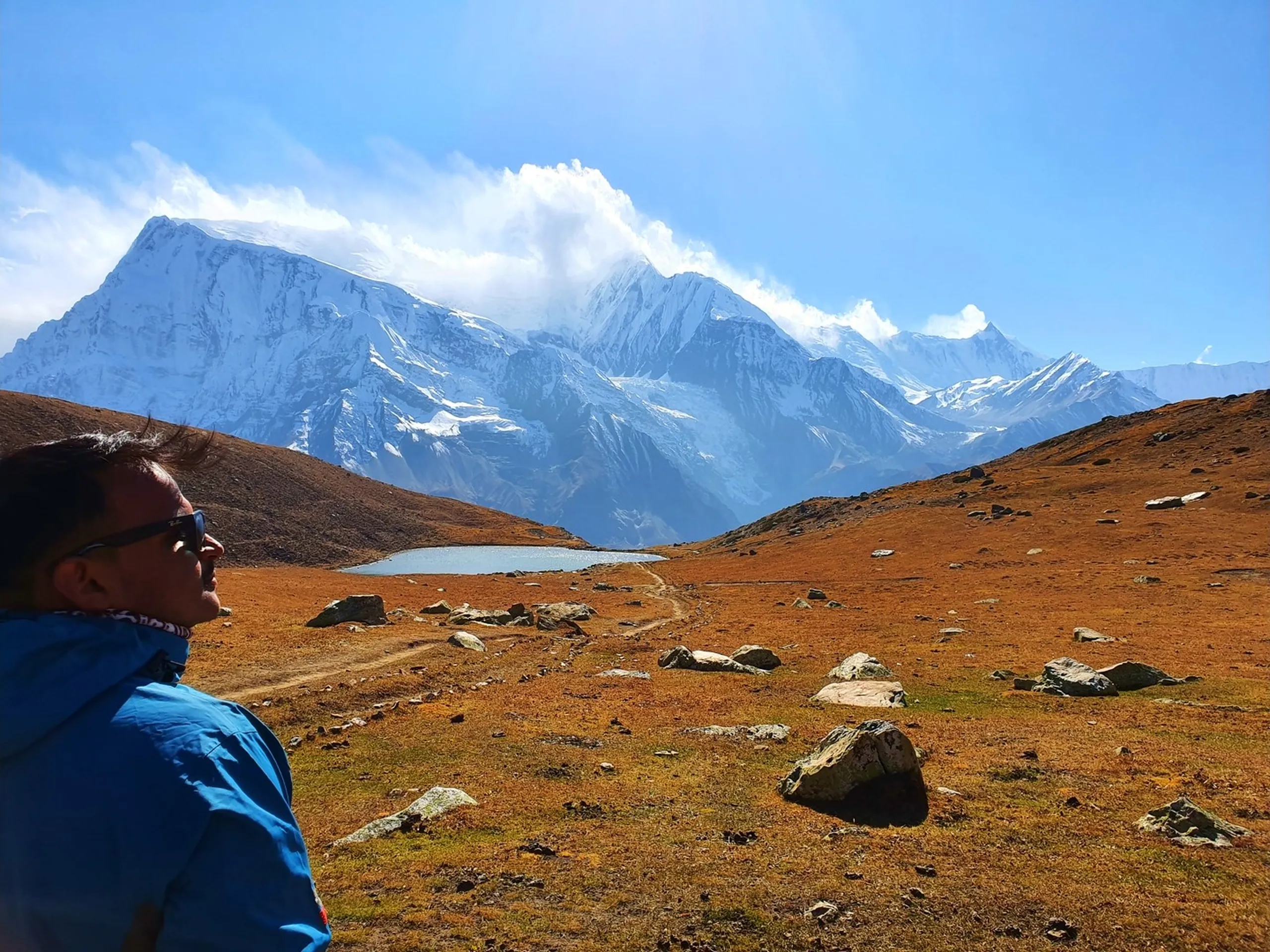 Side profile of trekker in blue Jack Wolfskin jacket with sunglasses gazing pensively at distant turquoise Ice Lake Manang Nepal from elevated viewpoint with vast panoramic Himalayan mountain range Gangapurna Annapurna massif covered in glacial snow golden autumn tundra moorland rocky landscape winding trail Annapurna Circuit Trek Nepal high altitude scenery travel photography