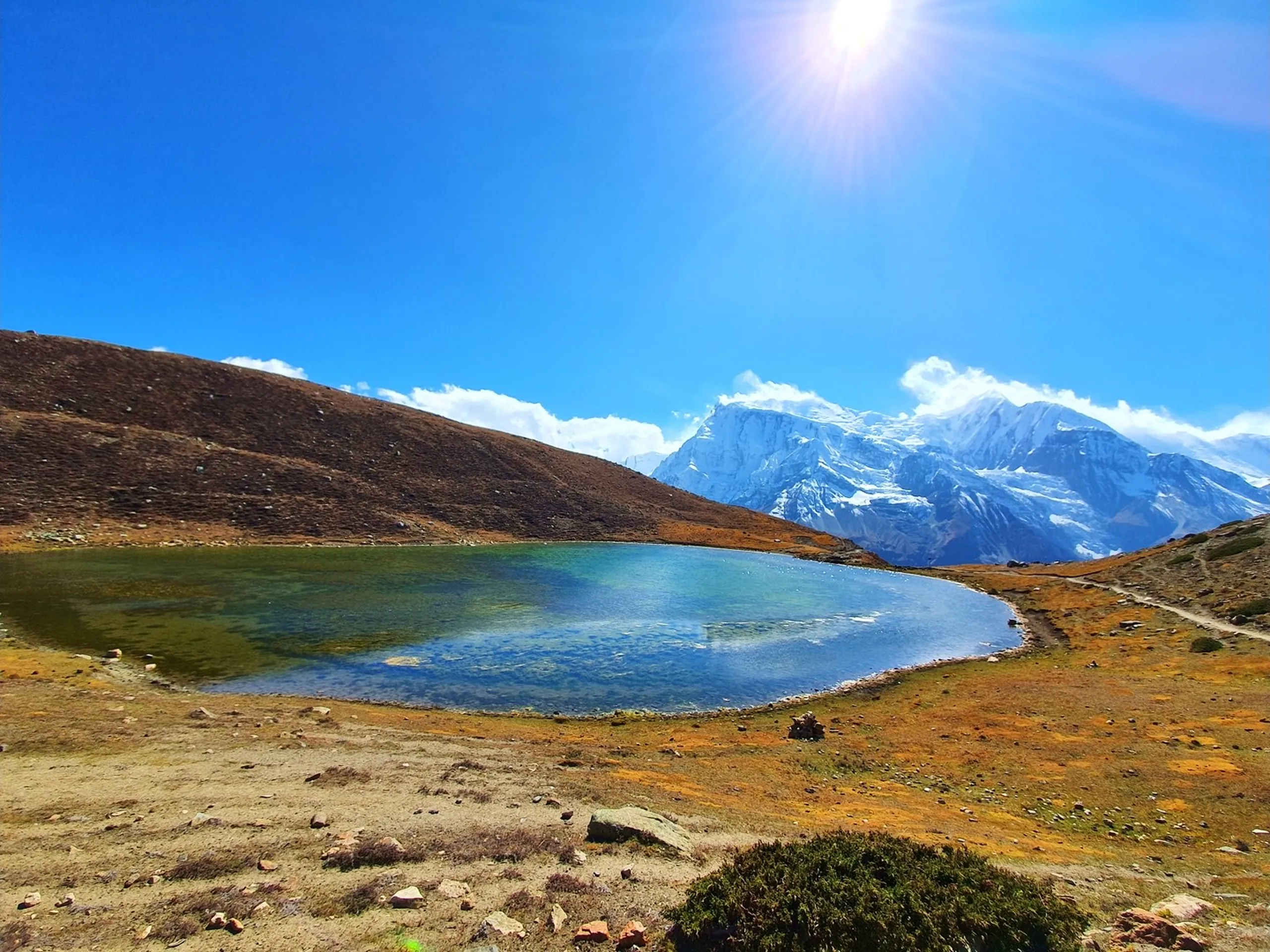 Pristine wide-angle landscape view of Ice Lake Manang Nepal 4600m altitude perfectly curved turquoise glacial alpine lake with brilliant sunburst lens flare in deep blue sky orange golden tundra meadow surrounding lake dramatic snow-capped Annapurna Himalayan peaks in background winding trail along lake shore Annapurna Circuit Trek Nepal must-visit destination travel photography