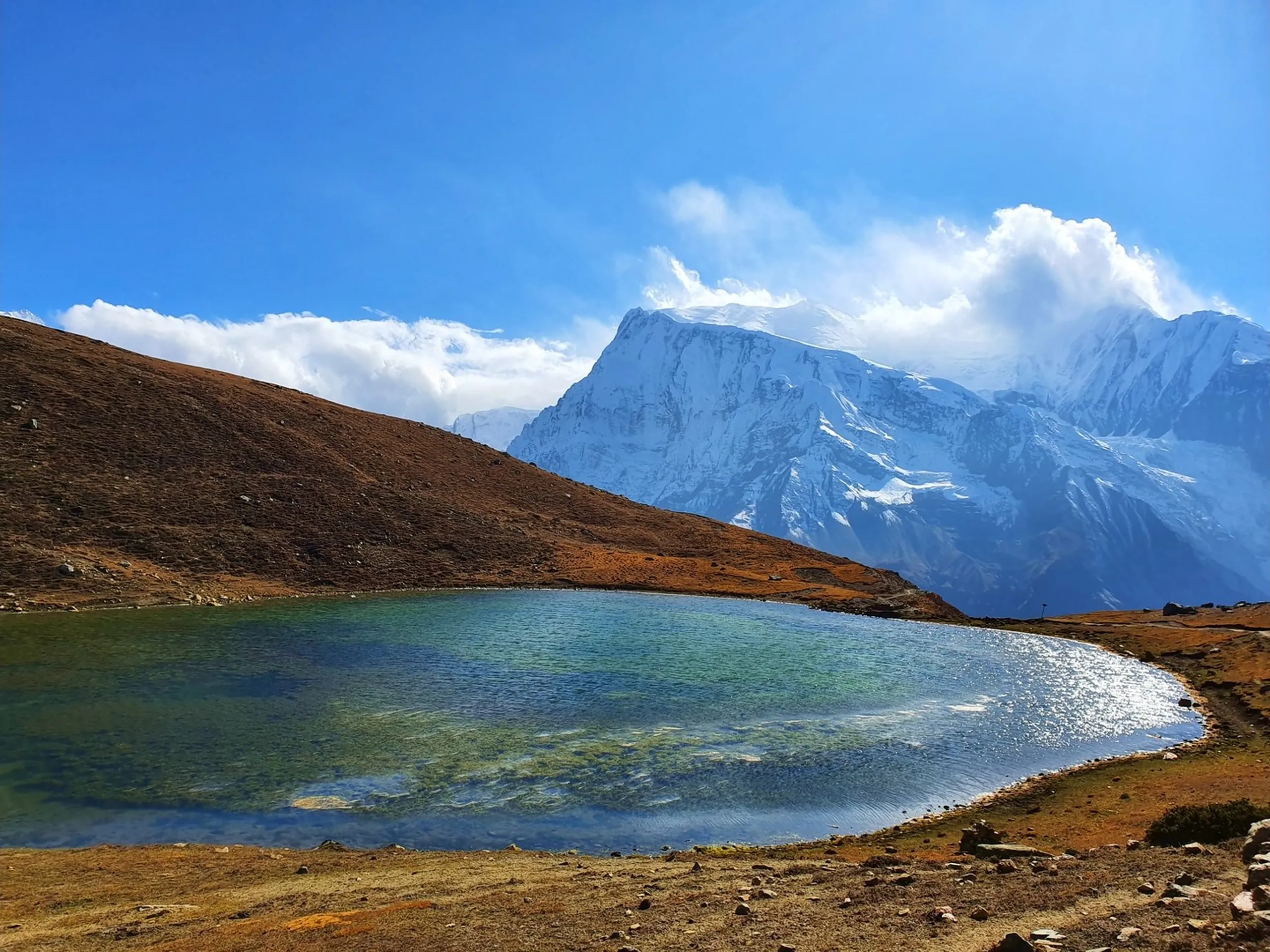 Stunning wide landscape photograph of Ice Lake Manang Nepal crystal clear turquoise glacial lake reflecting snow-capped Himalayan mountains Gangapurna peak with dramatic white cumulus clouds against deep blue sky orange and ochre high altitude tundra terrain in foreground Annapurna Circuit trek Nepal scenic viewpoint travel photography best lakes Nepal