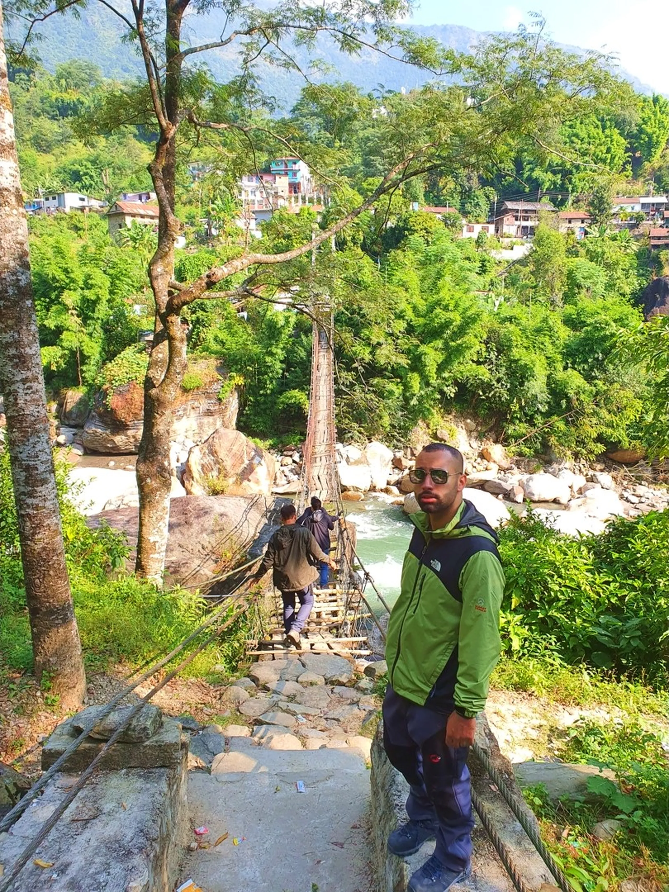 A young male trekker wearing a green North Face jacket, dark trekking pants, and aviator sunglasses stands confidently at the top of a rickety wooden suspension bridge over a rushing turquoise river in Nepal, posing for the camera while a companion in an olive jacket carefully crosses the swaying bridge behind him - dense jungle trees, massive boulders, and a small Nepali hillside village visible in the background - a thrilling Nepal suspension bridge crossing moment on the Annapurna Circuit trekking route, Besisahar, Lamjung