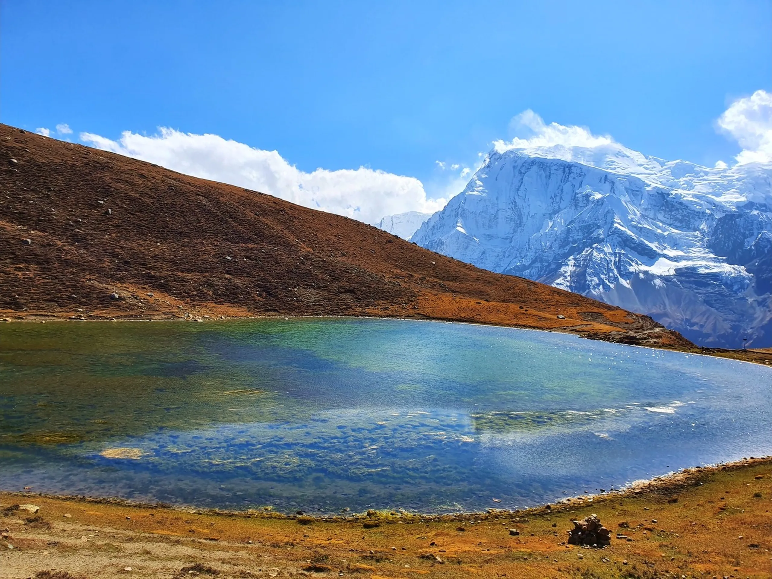 Serene close-up landscape of Ice Lake Manang Nepal transparent turquoise green glacial water revealing rocky lake floor massive snow-covered Himalayan mountain Gangapurna dominating skyline with white clouds golden brown alpine tundra surrounding shore in autumn Annapurna Circuit trek Nepal high altitude lake photography travel blog