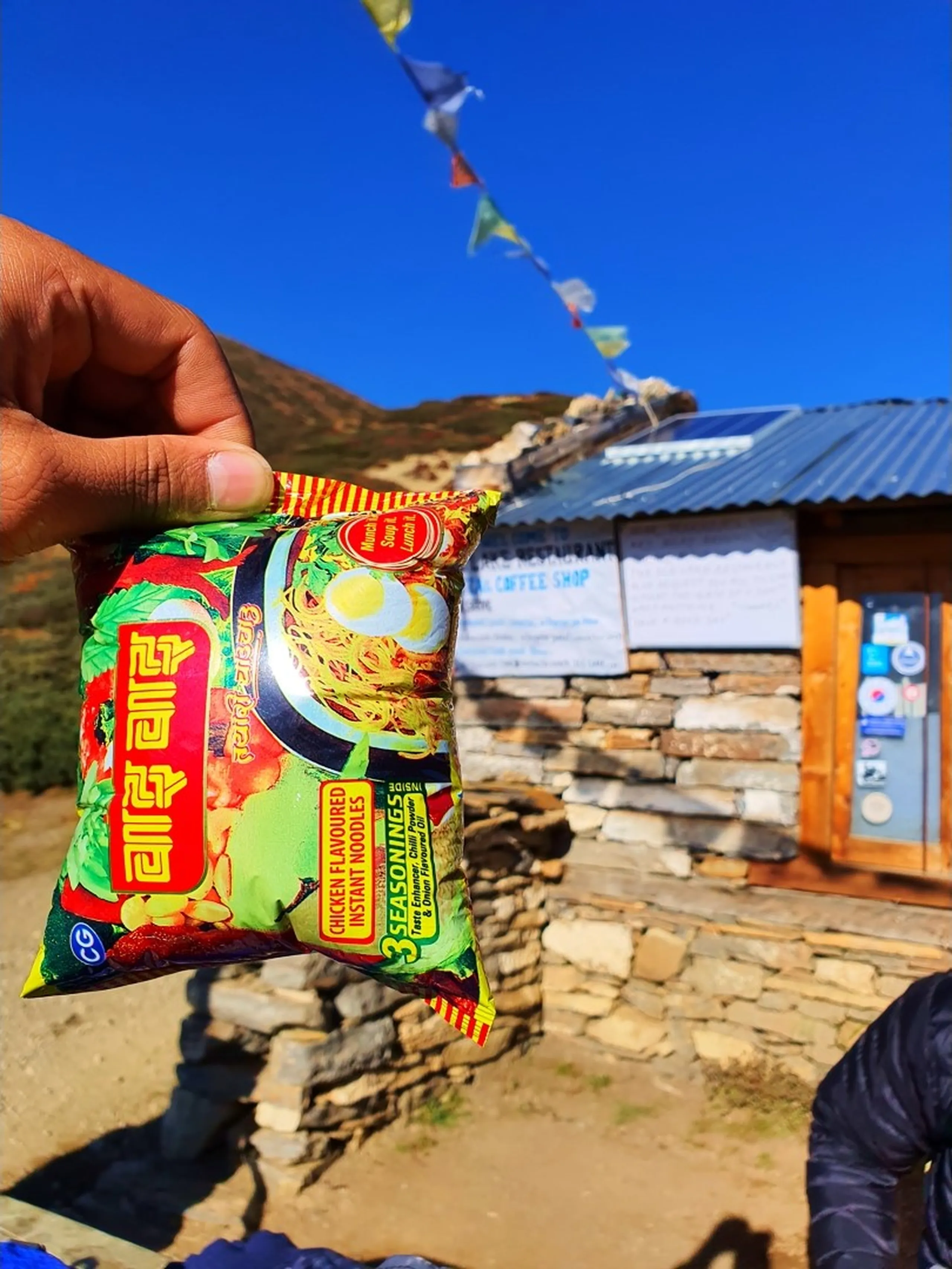 Hand holding a packet of Wai Wai chicken flavoured instant noodles Nepal's most iconic and beloved local snack brand against bright blue sky with colorful Buddhist prayer flags and traditional stone tea house coffee shop teahouse in background on Annapurna Circuit trek trail Manang Nepal trekking food snack culture travel blog Nepal local food