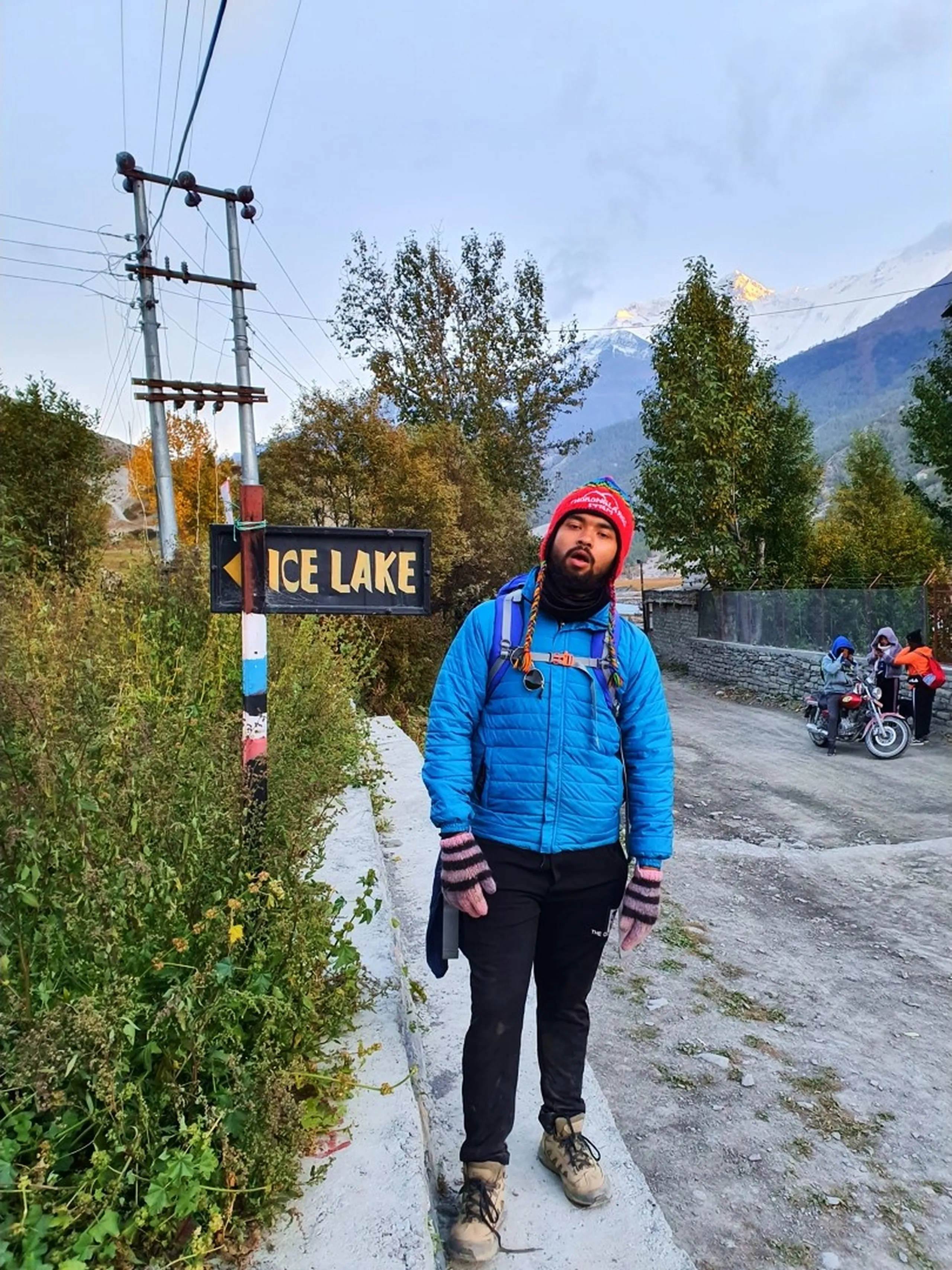 Exhausted but determined male trekker in blue puffer jacket with red Thamel knit beanie hat standing beside Ice Lake directional wooden signpost in Manang village Nepal at early morning with glowing Himalayan snow peaks in background autumn trees Annapurna Circuit trek Nepal trekking fatigue humor travel blog