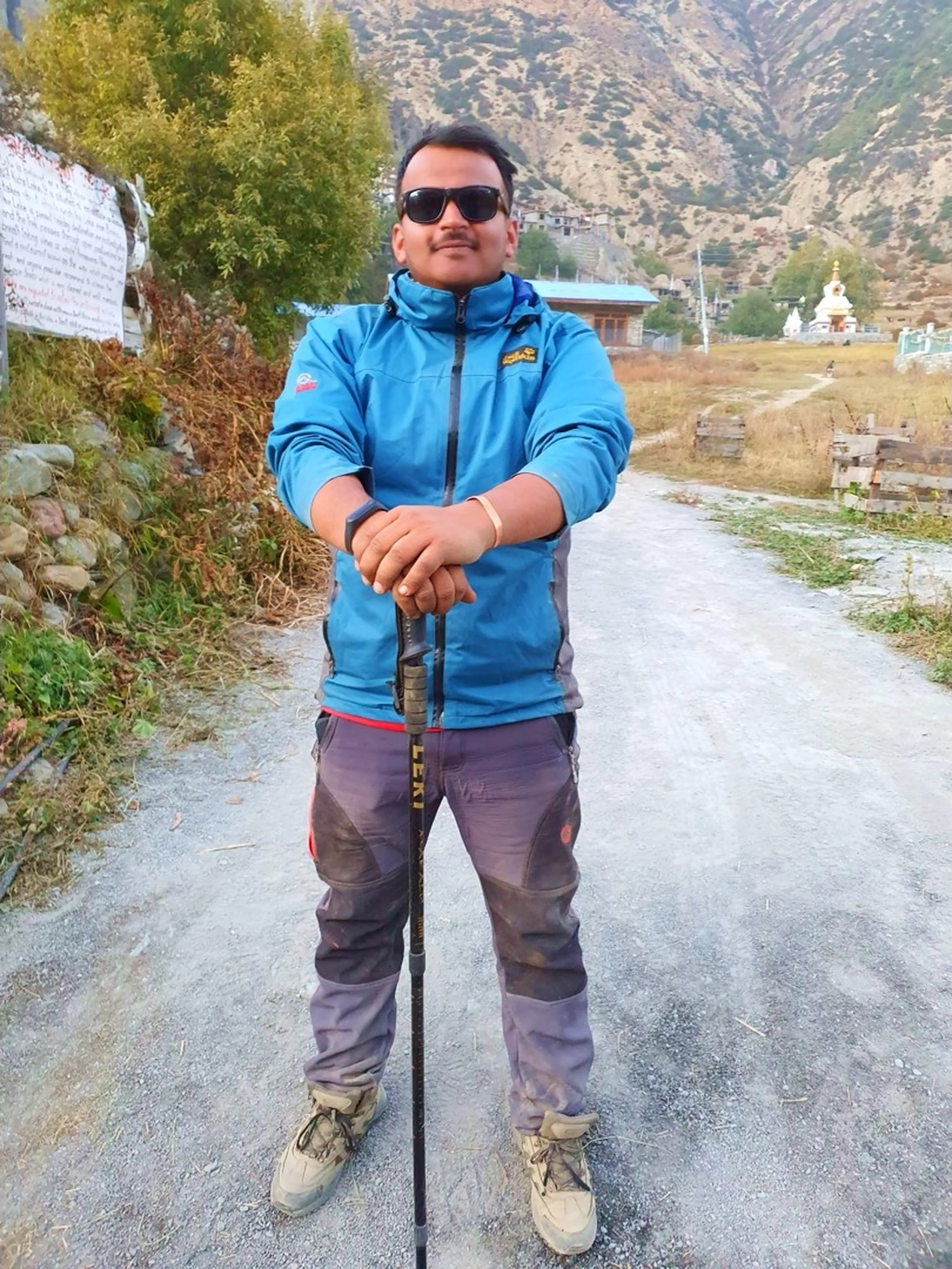Confident smiling male trekker in blue Jack Wolfskin waterproof jacket dark sunglasses standing proud on village pathway in Manang Nepal leaning on LEKI trekking pole with traditional whitewashed Buddhist stupa monastery visible in background after successful descent from Ice Lake 4600m Annapurna Circuit trek achievement Nepal adventure travel