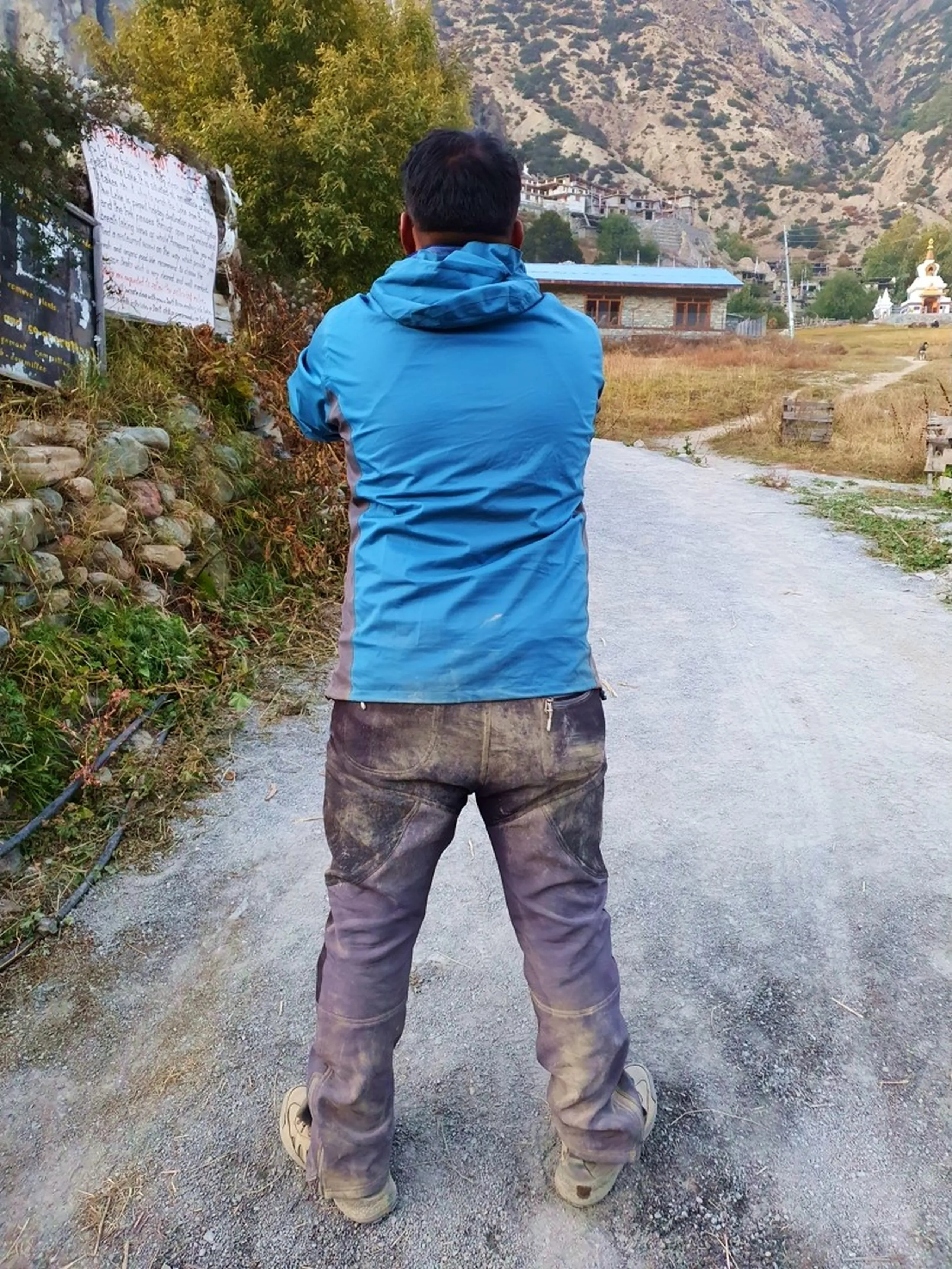 Rear view of male trekker in blue waterproof jacket walking away on stone pathway in Manang village Nepal with heavily mud-caked and dust-covered trekking pants legs showing evidence of difficult steep mountain descent from Ice Lake Annapurna Circuit trek mud trail conditions Nepal trekking reality travel photography authentic experience