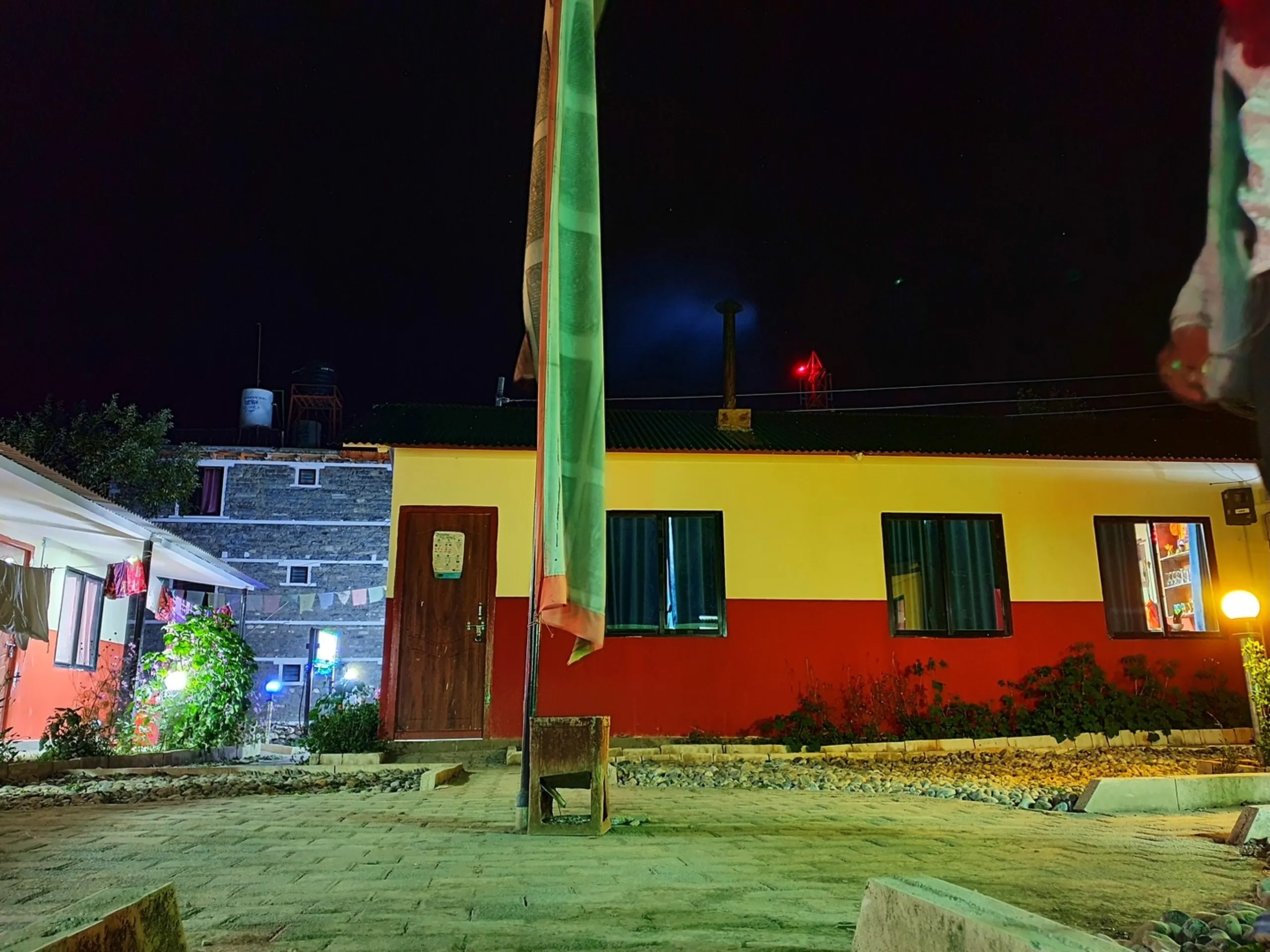 Nighttime exterior view of colorful guesthouse teahouse lodge in Manang Nepal painted bright yellow upper half and red lower half with traditional Buddhist prayer pole lung ta in foreground dark starry night sky warm glowing interior lights through curtained windows stone building neighboring guesthouse garden plants Annapurna Circuit trek accommodation Nepal budget travel lodging