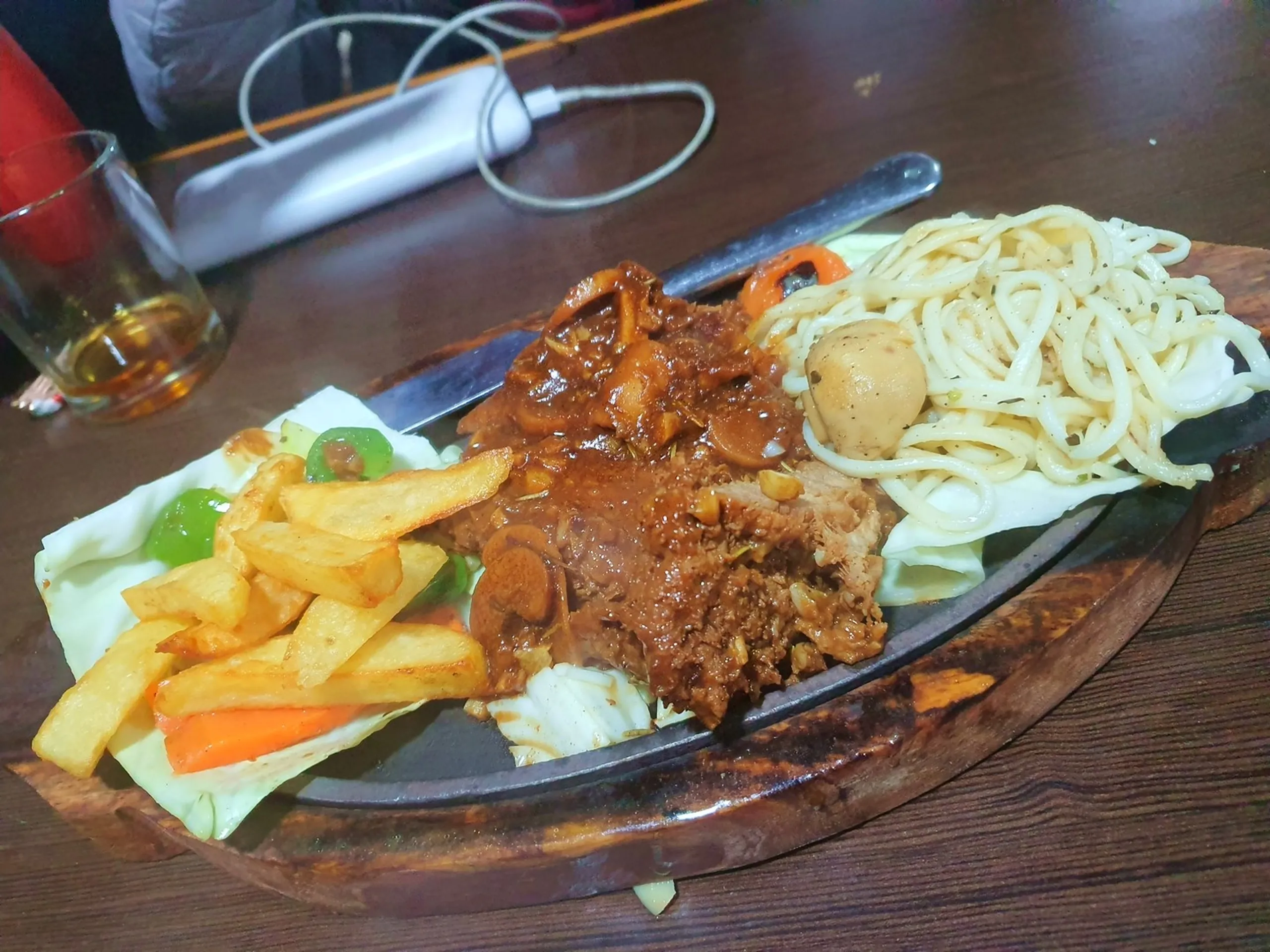 Sizzling hot yak steak served on traditional iron skillet wooden board at Alpine Home restaurant Manang Nepal garnished with French fries sauteed mushrooms in brown gravy spaghetti noodles with button mushrooms capsicum vegetables local Himalayan cuisine trekker dinner Annapurna Circuit Nepal mountain restaurant food travel blog must-try yak meat dish