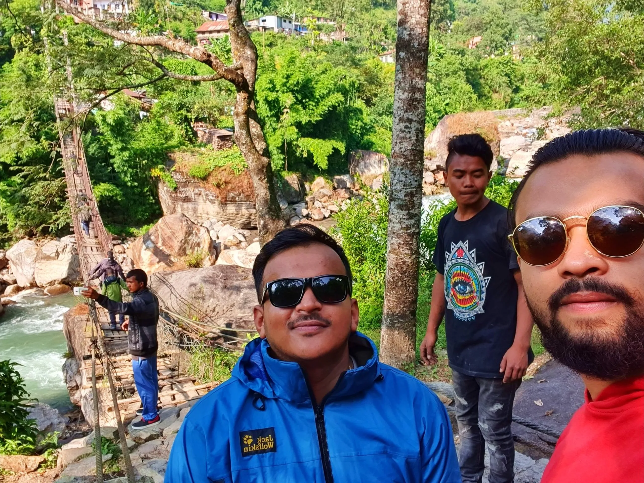 A close-up selfie of three young Nepali trekkers near a traditional rope suspension bridge over a fast-flowing mountain river in Nepal - one wearing a blue Jack Wolfskin jacket with black sunglasses smiling broadly in the center, another in a black graphic tee with a spiky hairstyle, and the third with round gold-framed sunglasses barely visible at the edge - another trekker visible balancing carefully on the rickety bridge in the background - lush green jungle and large river boulders framing the scene - a candid Nepal trekking friendship moment on the Annapurna Circuit route