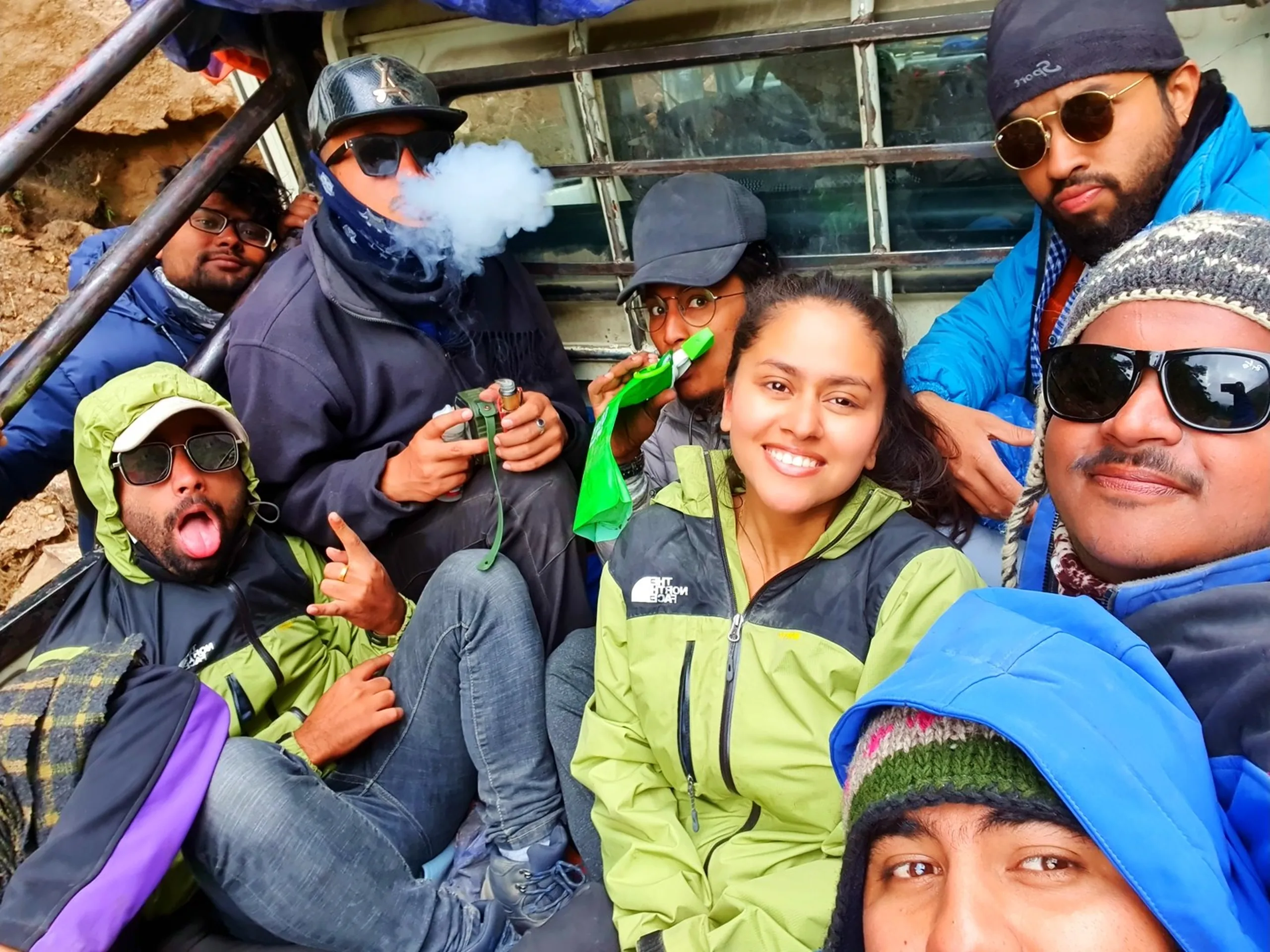 Lively group selfie of ten trekkers crammed together in the back of an open Bolero jeep on dusty mountain road returning from Annapurna Circuit trek Nepal one trekker in blue face mask blowing vape smoke cloud another in North Face lime green jacket smiling woman bearded man in grey knit hat sunglasses blue puffer jackets mixed trekking outerwear adventure travel group Nepal mountain jeep ride real trekking experience humor