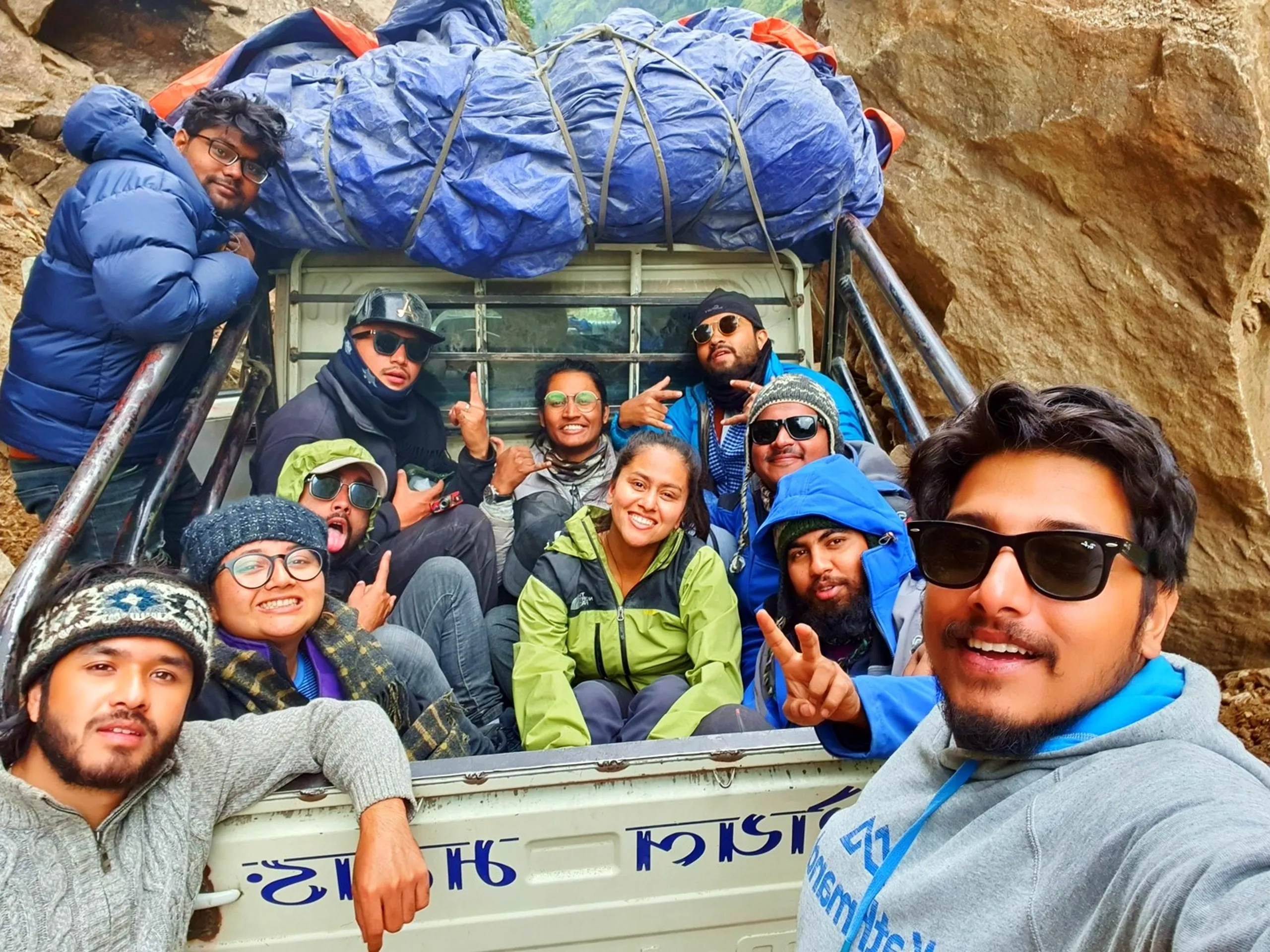 Wide group selfie of entire Annapurna Circuit trek squad packed into back of white Bolero jeep with massive blue tarpaulin-covered luggage pile strapped on roof rack with cargo net stopped at narrow rocky mountain pass Nepal trekkers making peace signs shaka gestures tongues out beaming smiles grey knit beanie North Face lime green jacket blue puffer jackets Nepali text on jeep tailgate authentic Nepal mountain transport group travel adventure