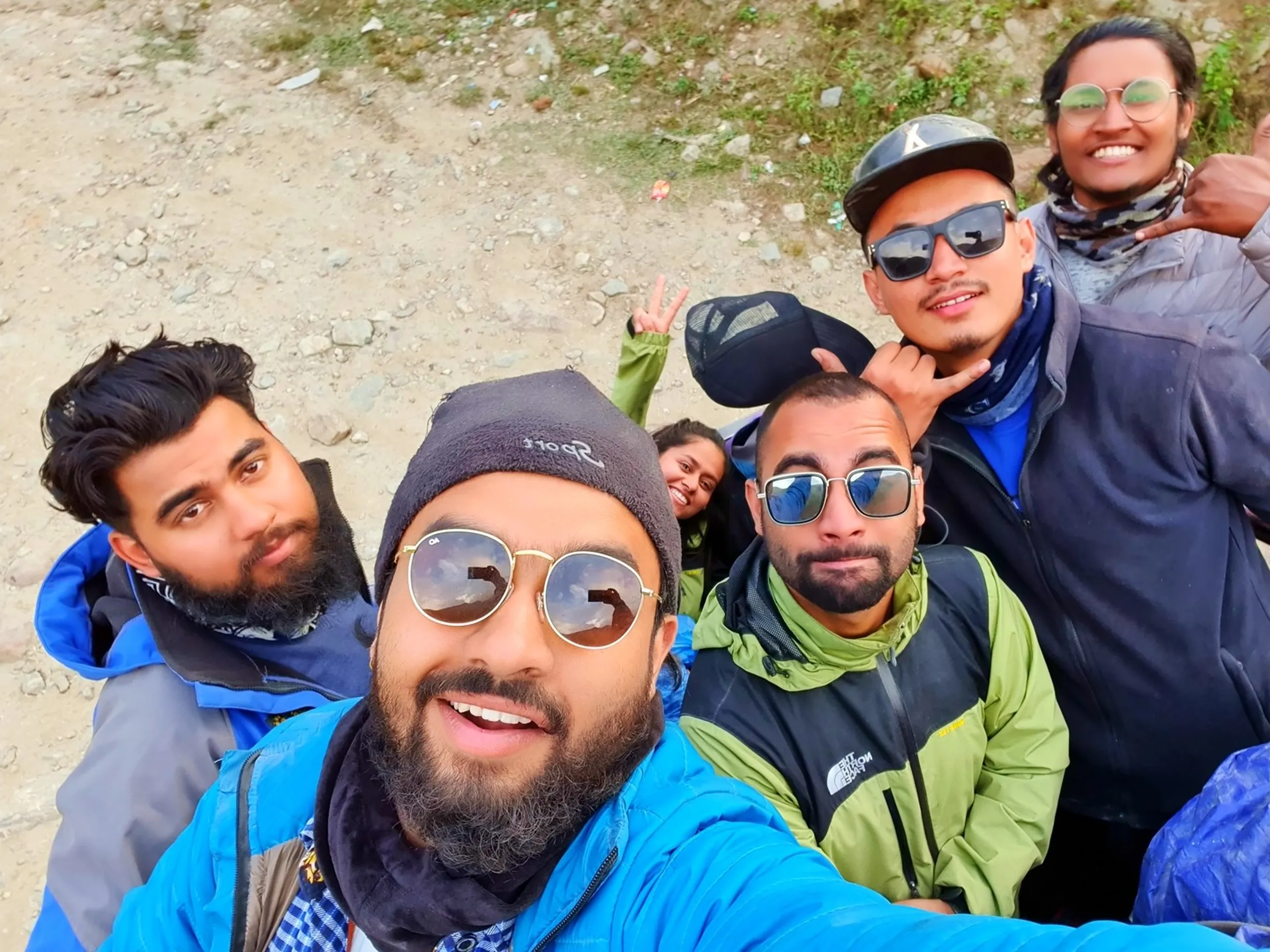 Close-up selfie of six smiling trekkers during stop on rough mountain road Nepal Annapurna region man in foreground with full beard round gold-frame mirror sunglasses blue jacket laughing mouth open bearded man beside him blue jacket blue puffer man with reflective sunglasses dark cap two men in background one in North Face lime green jacket dark cap sunglasses smiling woman in green jacket peeking over shoulder Nepal group travel adventure trekking friends