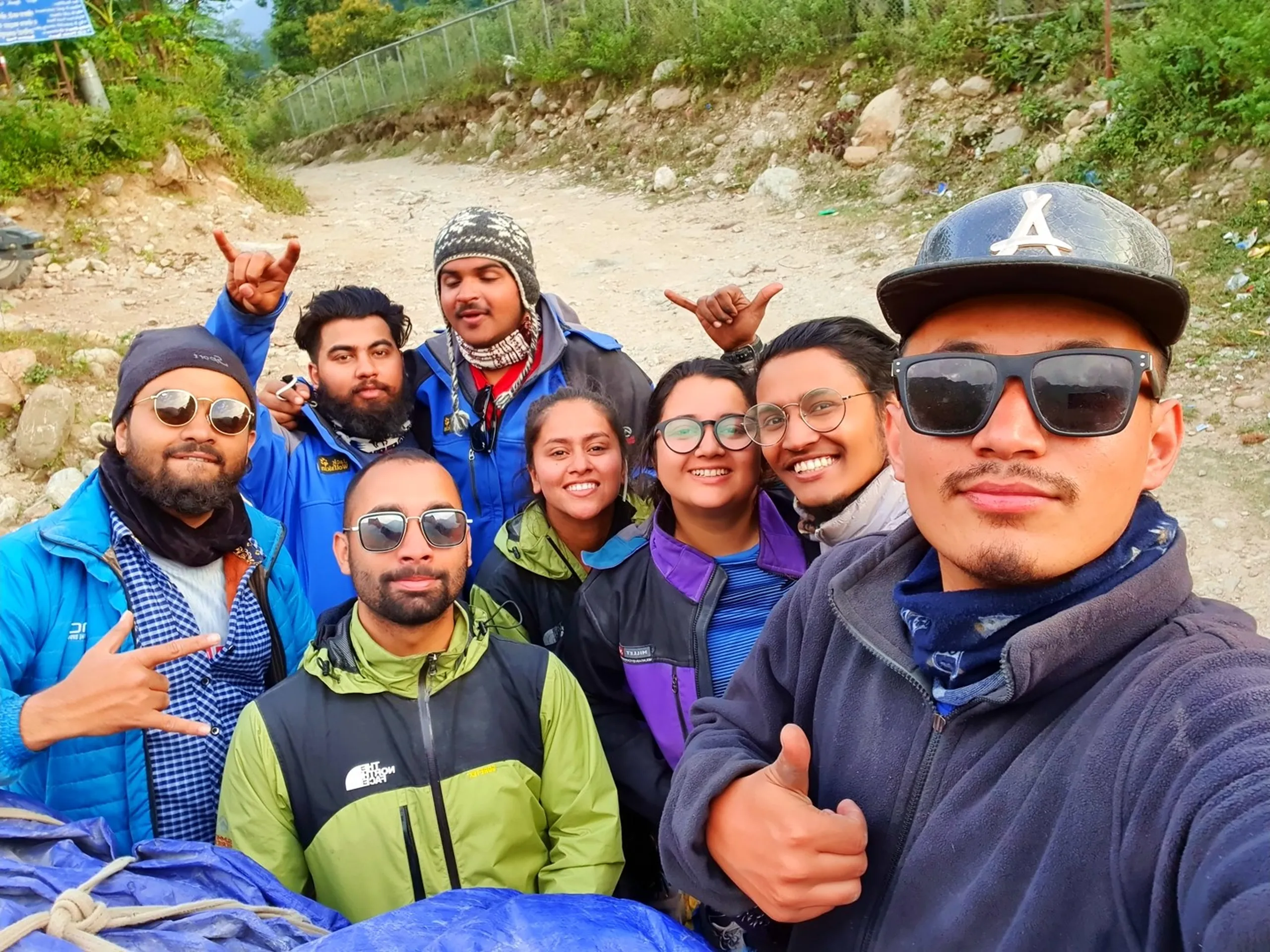 Group of eight trekkers posing together during road stop on gravel mountain highway Nepal hills Annapurna region lush green vegetation and signboard visible in background diverse mixed group men and women in trekking gear North Face lime green jacket Jack Wolfskin blue jacket knit grey beanie red scarf blue puffer jacket purple jacket round glasses shaka and pointing gestures all smiling Nepal Annapurna Circuit trek group return journey Besisahar Lamjung