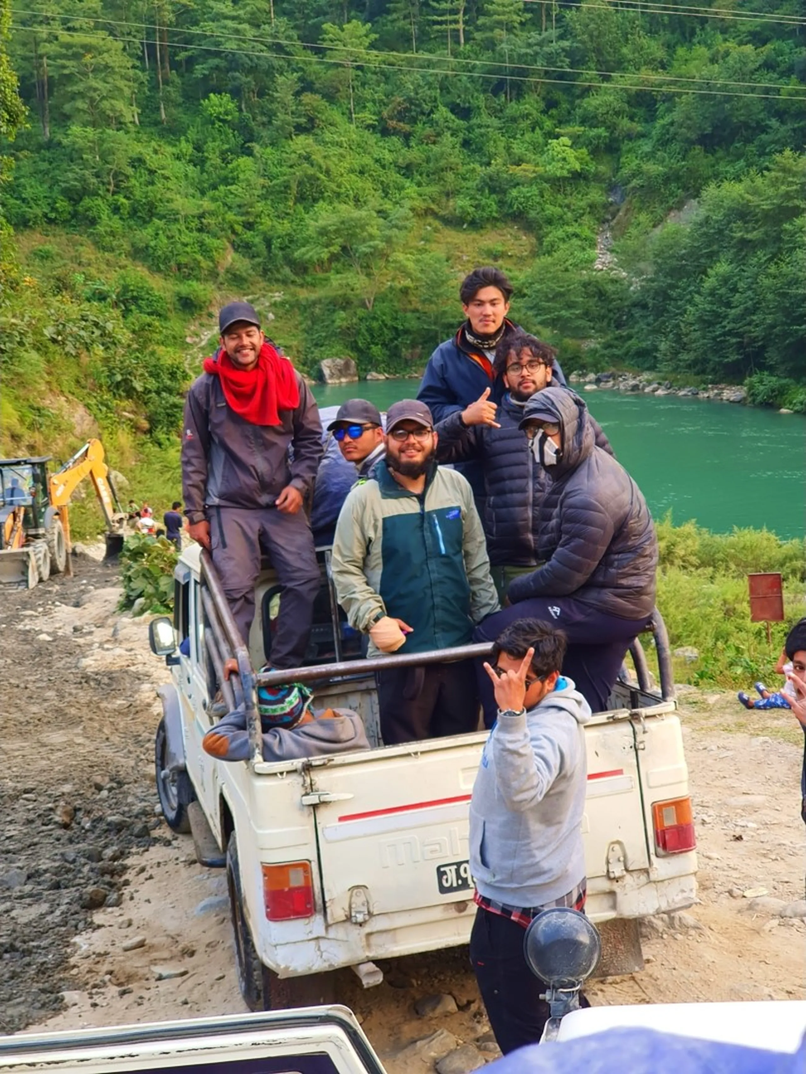 Six young male trekkers crowded in the back of a white Mahindra jeep on a narrow mountain road Nepal lush dense green pine forested hillside and vivid turquoise Marsyangdi River gorge visible in background man in red scarf standing tall smiling others seated showing thumbs up and rock-on gestures blue puffer jacket grey hoodie dark jacket Annapurna Circuit trek Nepal adventure travel return journey river valley