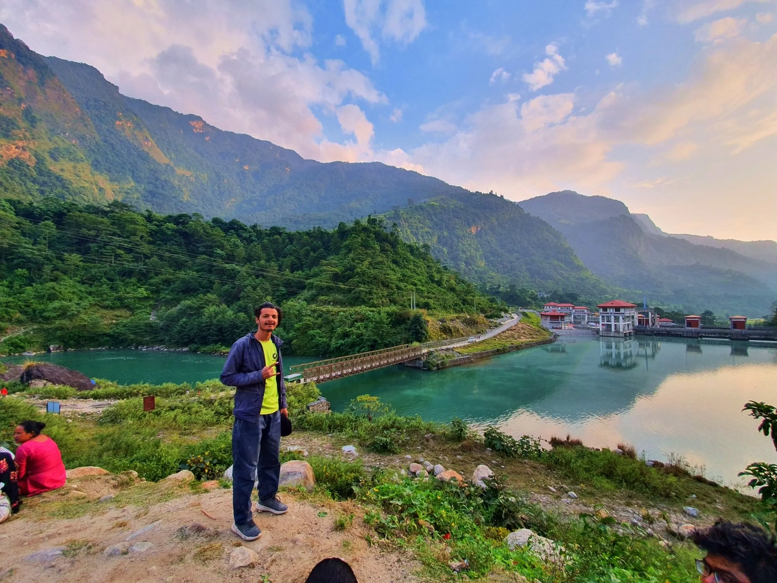 Young male trekker in navy blue jacket and green t-shirt smiling and gesturing with finger gun pose at Khudi Hydropower Dam viewpoint Besisahar Lamjung Nepal with stunning panoramic view of luminous turquoise Marsyangdi River reflecting golden evening light dense green forested Himalayan hills and pink-orange sunset cloudy sky suspension footbridge dam building Annapurna Circuit trek Nepal scenic stop travel photography