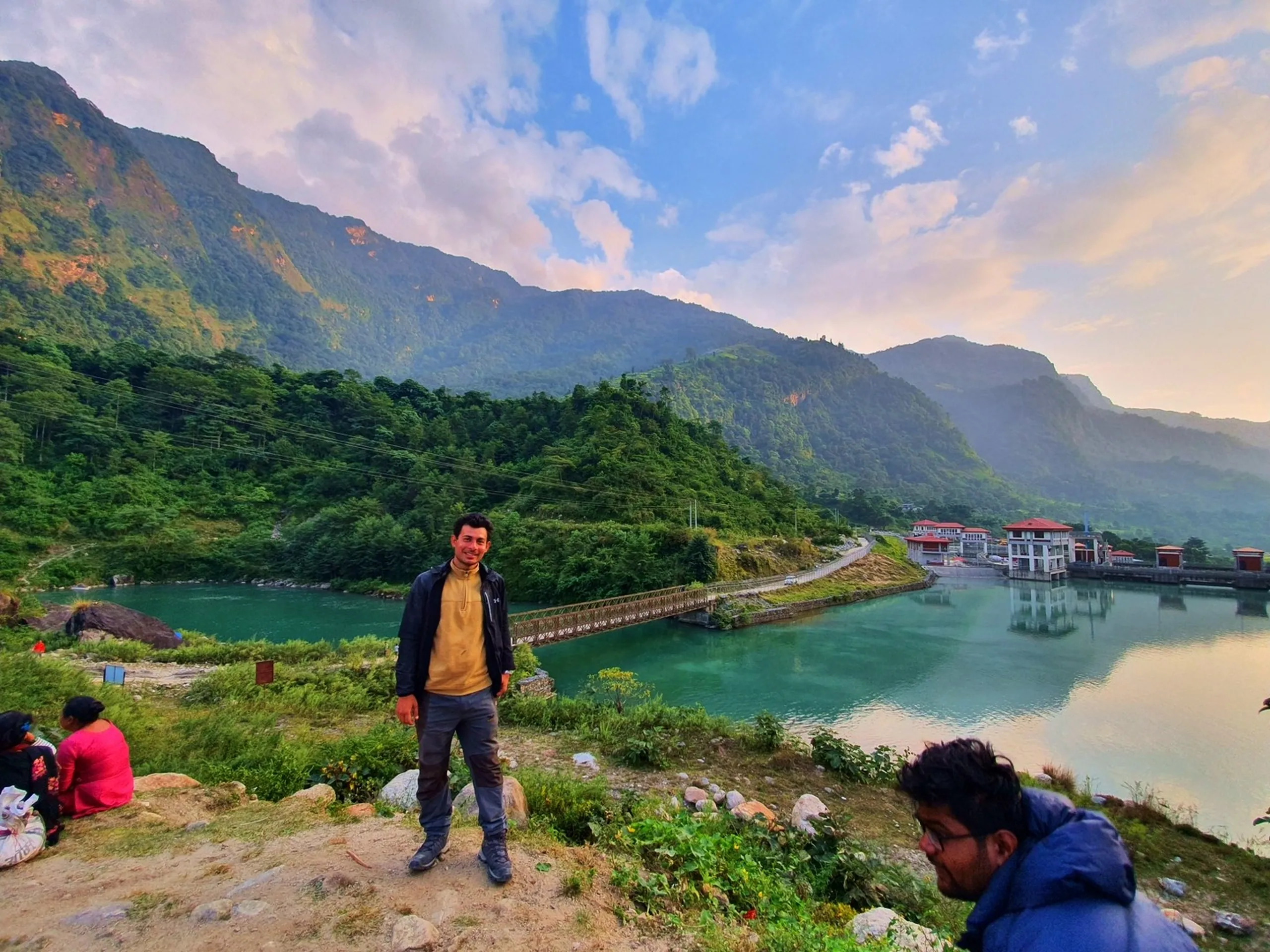 Smiling male trekker in mustard yellow hoodie and black waterproof jacket standing at elevated viewpoint overlooking Khudi Hydropower Dam Besisahar Lamjung Nepal with brilliant turquoise Marsyangdi River valley dense green forested Himalayan foothills suspension bridge dam building pink sunset clouds gorgeous Nepal river valley scenery Annapurna Circuit trek return journey travel photography