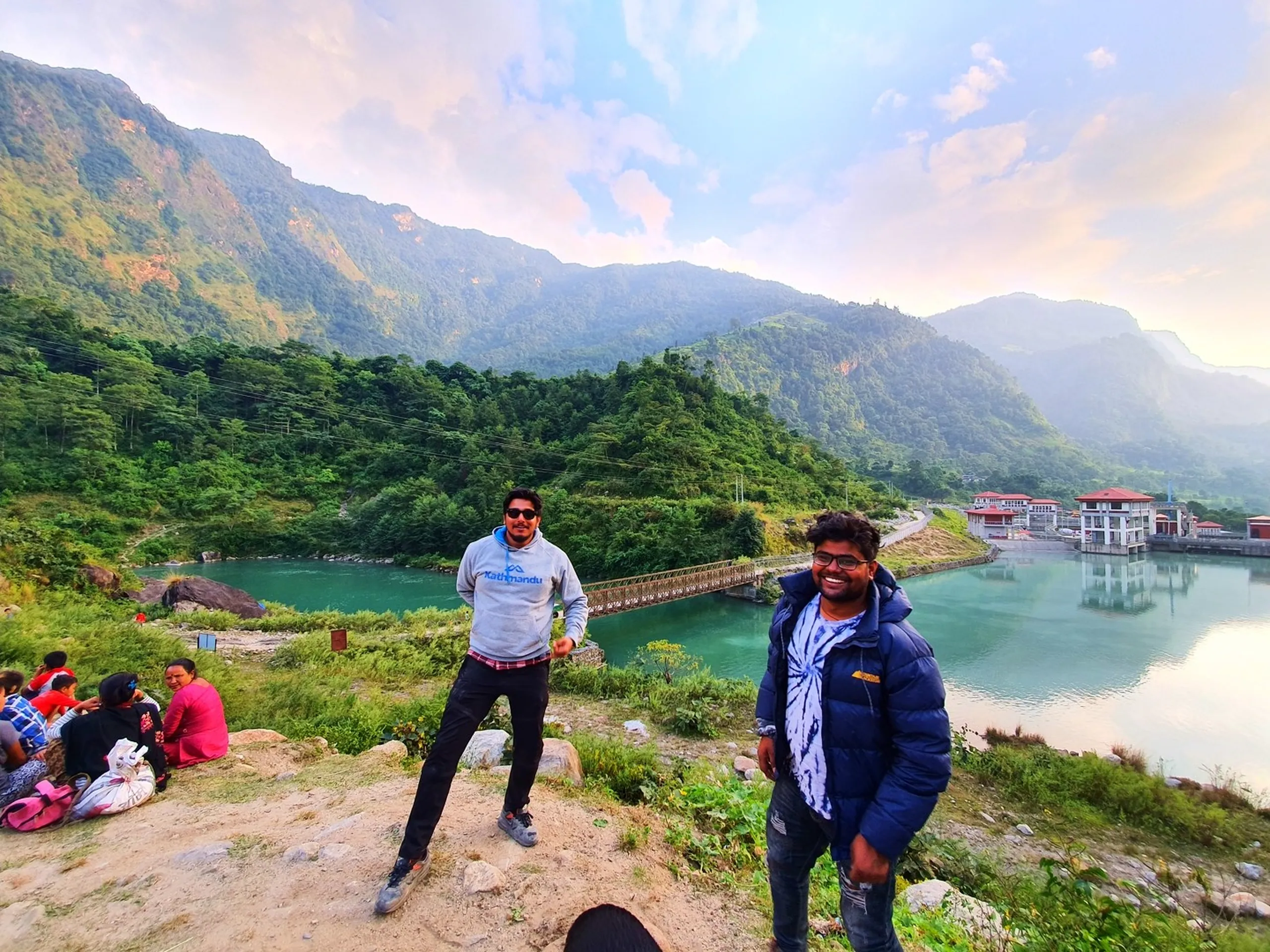 Two male trekkers posing together at Khudi Hydropower Dam scenic overlook Besisahar Lamjung Nepal at golden hour one wearing grey Kathmandu brand hoodie sunglasses other in white tie-dye shirt navy puffer jacket both smiling lush turquoise Marsyangdi River valley below green forested Himalayan hills suspension footbridge dam facility sunset pink clouds Nepal travel photography Annapurna Circuit trek friends