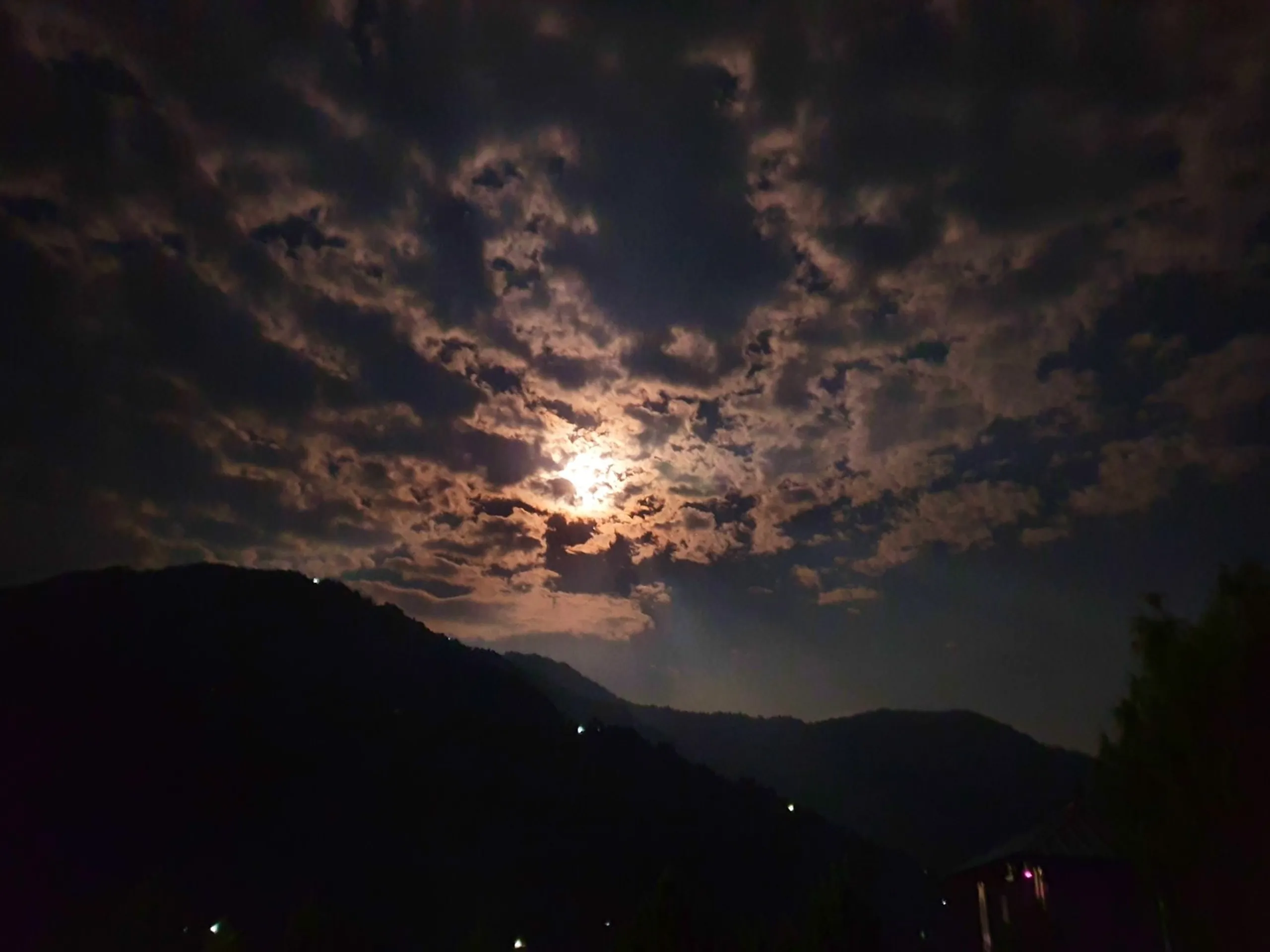 Dramatic atmospheric night sky photograph taken at Gateway Himalaya Resort Besisahar Lamjung Nepal showing full moon partially obscured by dramatic thick scattered cloud formations moonlight illuminating clouds in warm amber and silver tones dark silhouette of Himalayan foothills and tree in foreground scattered lights of village below moody Nepal night sky mountain travel photography