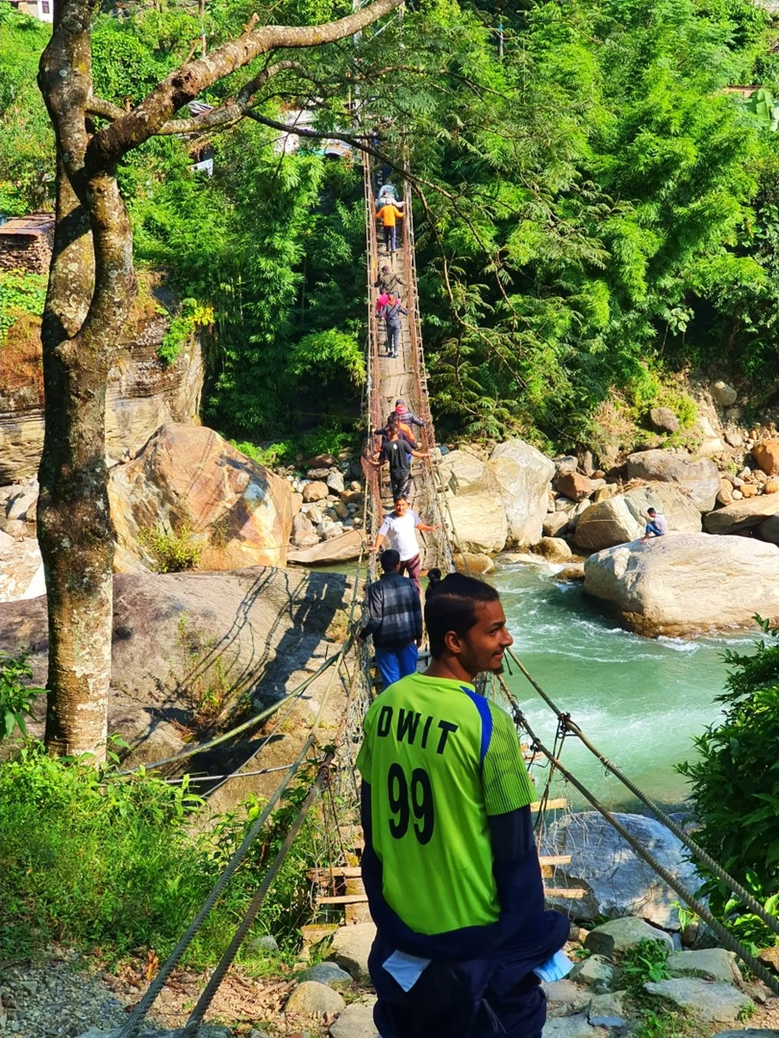A wide-angle shot of a group of young Nepal trekkers crossing a narrow, rope-and-plank suspension bridge over a clear turquoise mountain river - a young man in a neon green jersey numbered 99 stands at the near end watching as five others make their way across the swaying bridge one by one - giant mossy boulders, white water rapids, and dense jungle canopy frame the scene - a dramatic and adventurous Nepal hiking bridge crossing photograph on the Annapurna Circuit trail near Besisahar, Lamjung, ideal for Nepal adventure trekking blog content