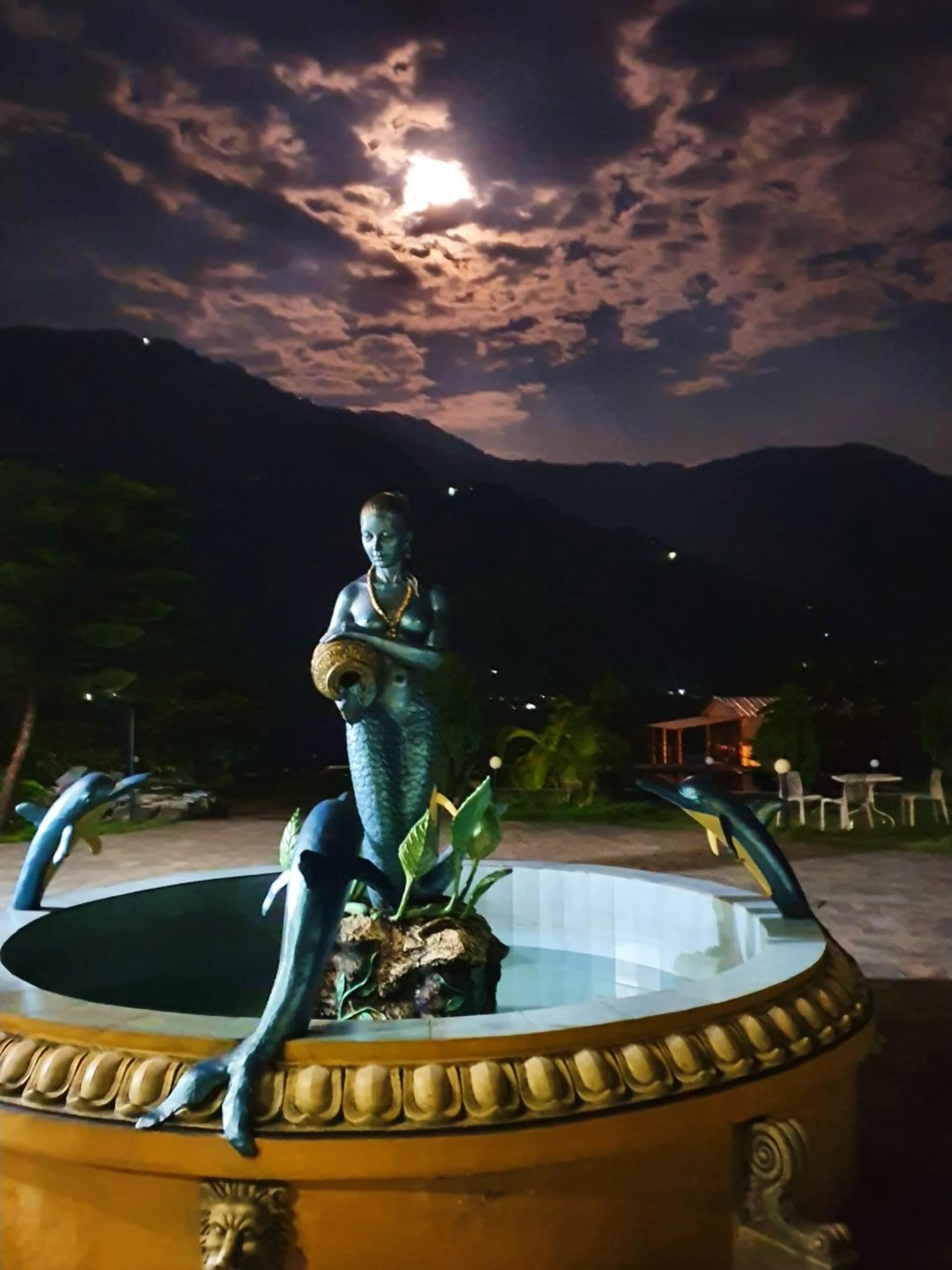 Atmospheric night photograph of decorative blue mermaid fountain statue with dolphin figures illuminated at Gateway Himalaya Resort Besisahar Lamjung Nepal dramatic full moon breaking through thick dark clouds above dark silhouette of Himalayan foothills garden seating area with warm yellow light visible in background Nepal luxury resort night photography travel blog