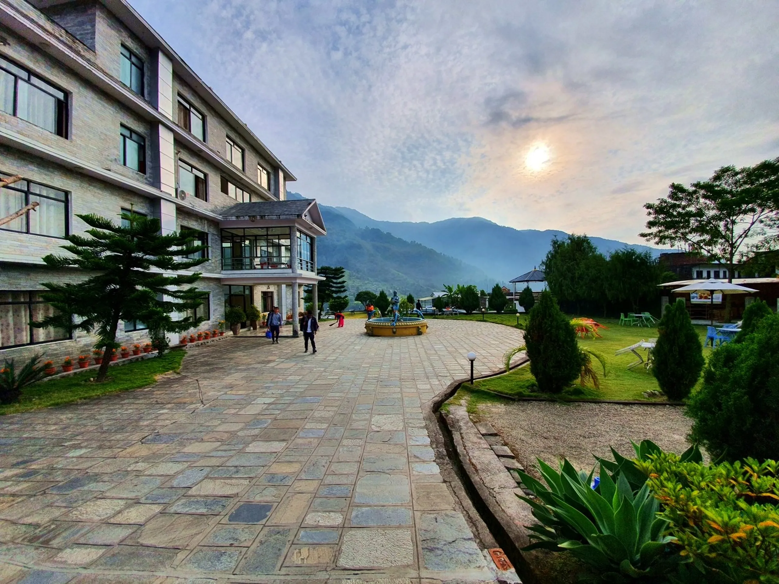 Wide angle early morning exterior view of Gateway Himalaya Resort hotel main building Besisahar Lamjung Nepal three-storey stone facade modern architecture large glass windows paved stone courtyard blue mermaid fountain centerpiece manicured garden lawns trimmed trees orange marigold flower pots misty Himalayan foothills in background overcast morning sky trekkers visible walking Nepal mountain resort accommodation travel photography