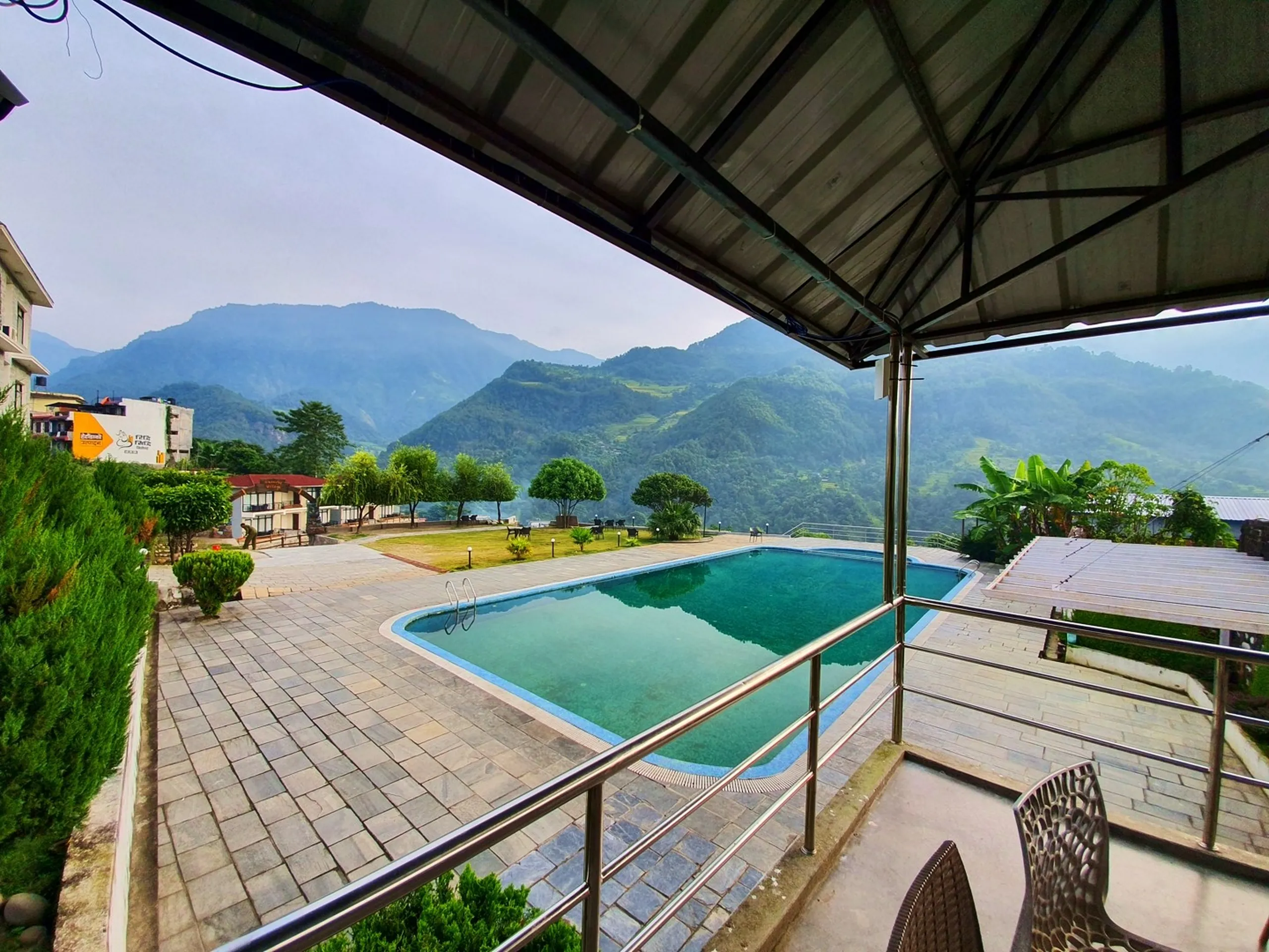 Elevated view from covered balcony terrace of Gateway Himalaya Resort Besisahar Lamjung Nepal showing rectangular turquoise swimming pool surrounded by paved stone deck manicured gardens lush green lawns Himalayan foothills layers of misty mountains in background overcast morning sky stainless steel railing in foreground Nepal mountain resort amenities pool view travel blog accommodation