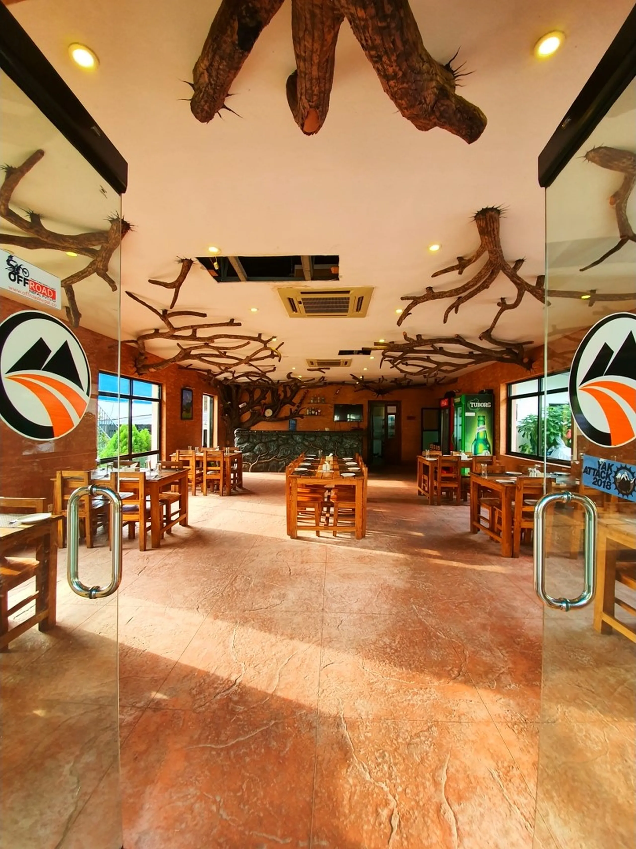 Interior of unique adventure-themed restaurant at Gateway Himalaya Resort Besisahar Lamjung Nepal featuring dramatic ceiling decoration of large natural tree trunk and branches spreading across white ceiling wooden dining tables and chairs polished terracotta tiled floor large glass windows mountain view Offroad Nepal logo sticker Tuborg beer fridge Yak Attack 2018 sticker Nepal trekker restaurant rustic decor dining travel