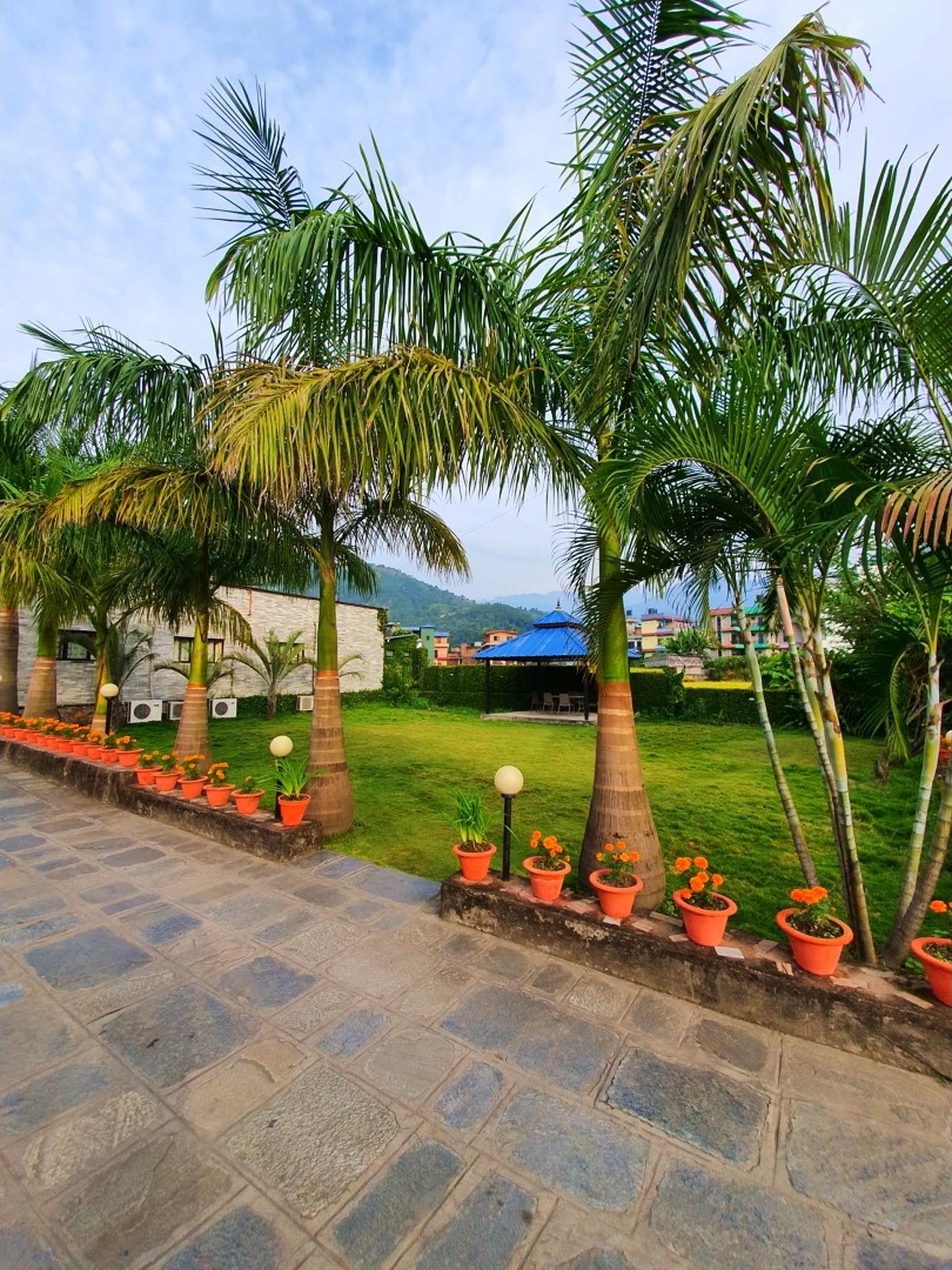 Lush tropical garden pathway at Gateway Himalaya Resort Besisahar Lamjung Nepal lined with tall elegant royal palm trees orange terracotta flower pots filled with orange marigolds globe path lights paved stone walkway lush green manicured lawn blue gazebo pavilion in background green Himalayan hills and partly cloudy sky Nepal mountain resort garden landscaping travel accommodation photography