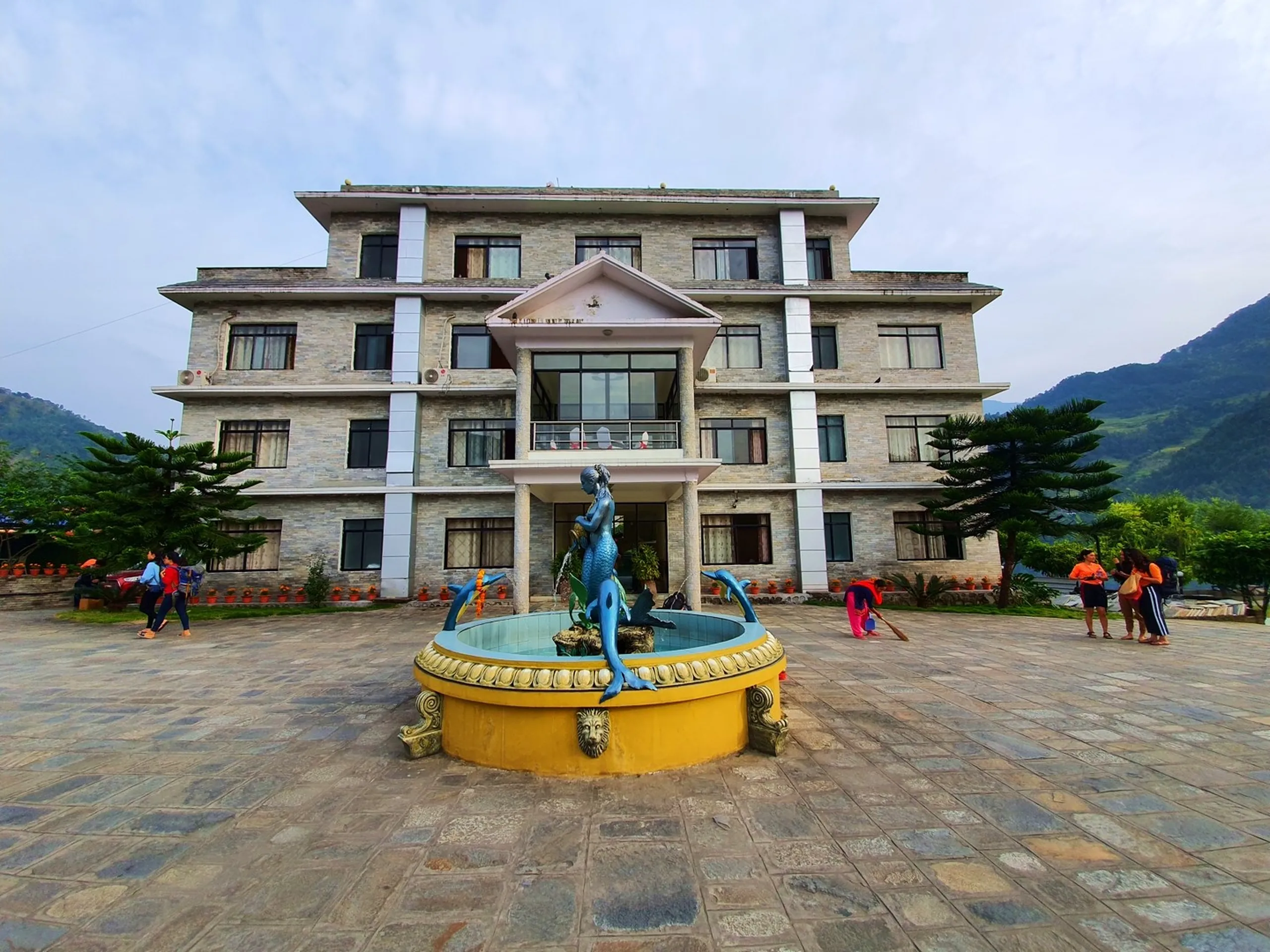 Full front exterior view of Gateway Himalaya Resort main building Besisahar Lamjung Nepal four-storey stone-clad facade with large glass balcony windows blue mermaid water fountain sculpture centerpiece on stone courtyard entrance guests visible walking lion-head base golden fountain detail trimmed pine trees orange marigold pots green Himalayan hills partly cloudy sky Nepal mountain hotel travel blog accommodation