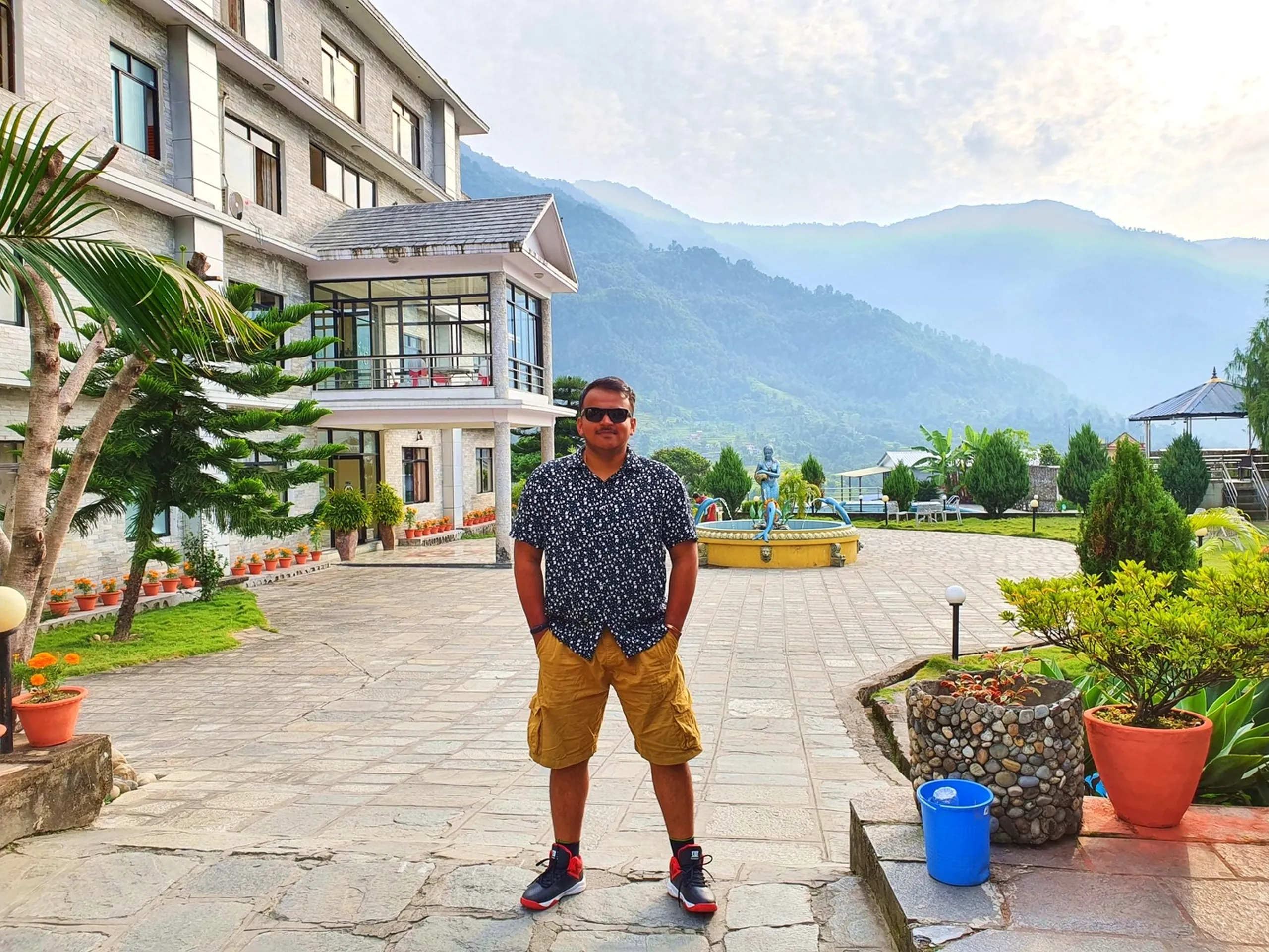 Male trekker wearing dark printed short-sleeve shirt khaki cargo shorts red-black Nike sneakers and dark sunglasses standing relaxed with hands in pockets in Gateway Himalaya Resort stone courtyard Besisahar Lamjung Nepal mermaid fountain behind him trimmed garden trees marigold pots resort main building visible misty green Himalayan hills background Nepal resort rest day after Annapurna Circuit trek adventure travel