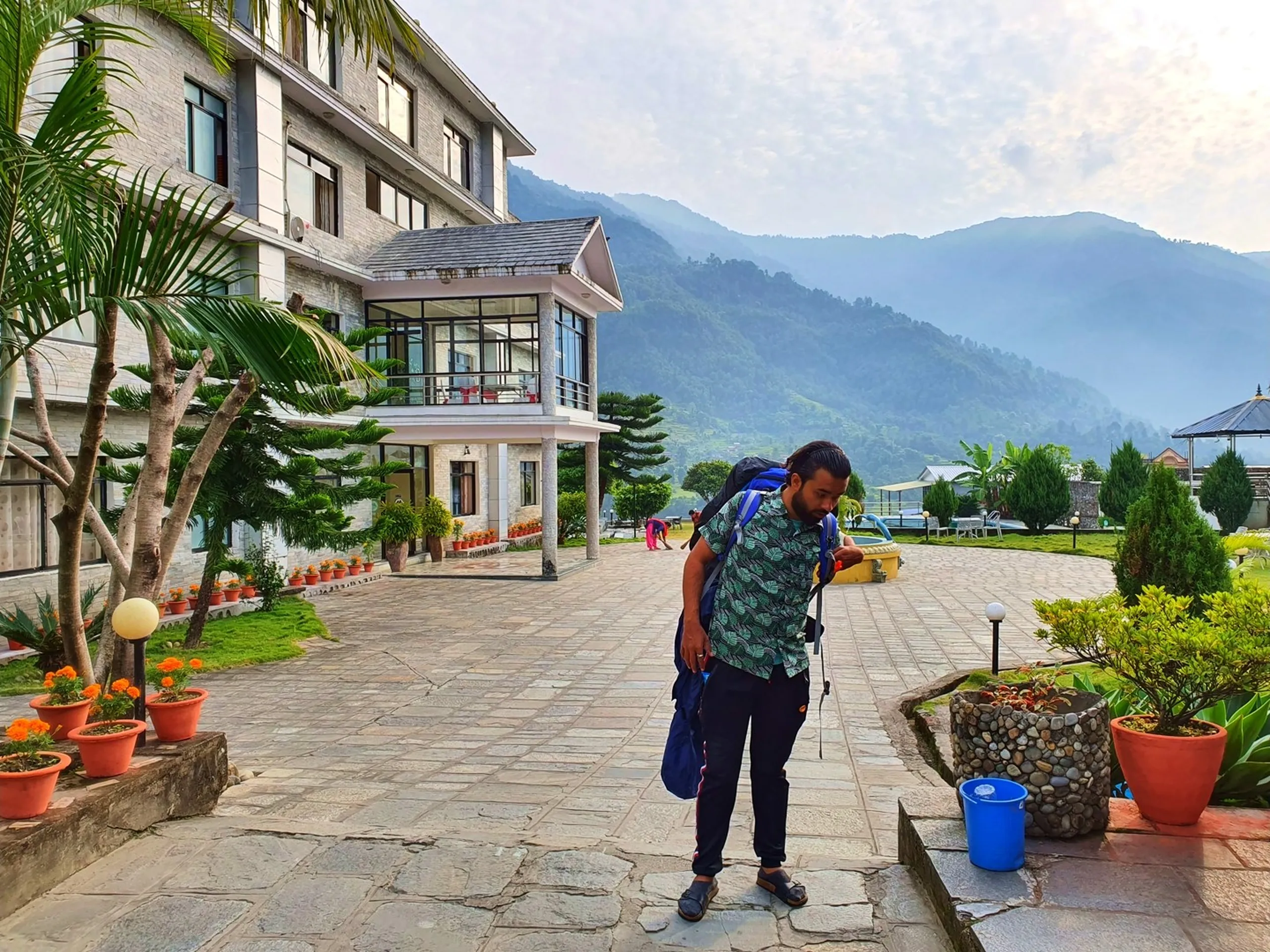 Male trekker in green tropical print shirt with large blue trekking backpack and trekking pole bending over adjusting gear on stone paved courtyard of Gateway Himalaya Resort Besisahar Lamjung Nepal garden palm trees marigold pots resort facade misty green Himalayan foothills visible heading back to bus Pokhara Annapurna Circuit trek departure Nepal travel blog packing up morning checkout