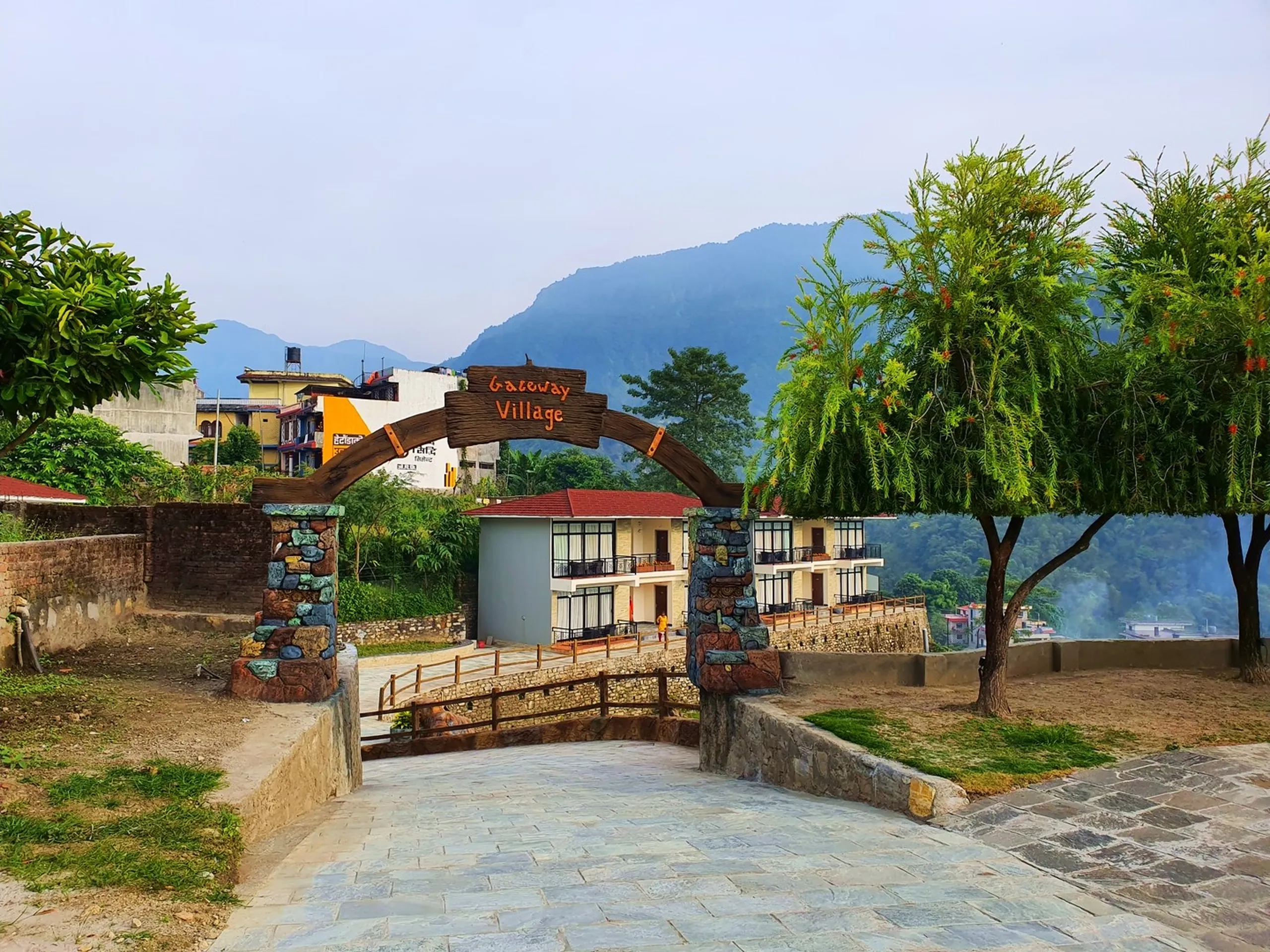 Rustic wooden arch entrance gate of Gateway Village resort complex Besisahar Lamjung Nepal with orange hand-lettered carved wooden sign reading Gateway Village stone pillar gateposts stone paved pathway modern multi-storey balcony guesthouse building visible through arch lush trees green lawn Himalayan foothills in morning mist Nepal mountain resort entrance gateway travel blog Annapurna Circuit starting point