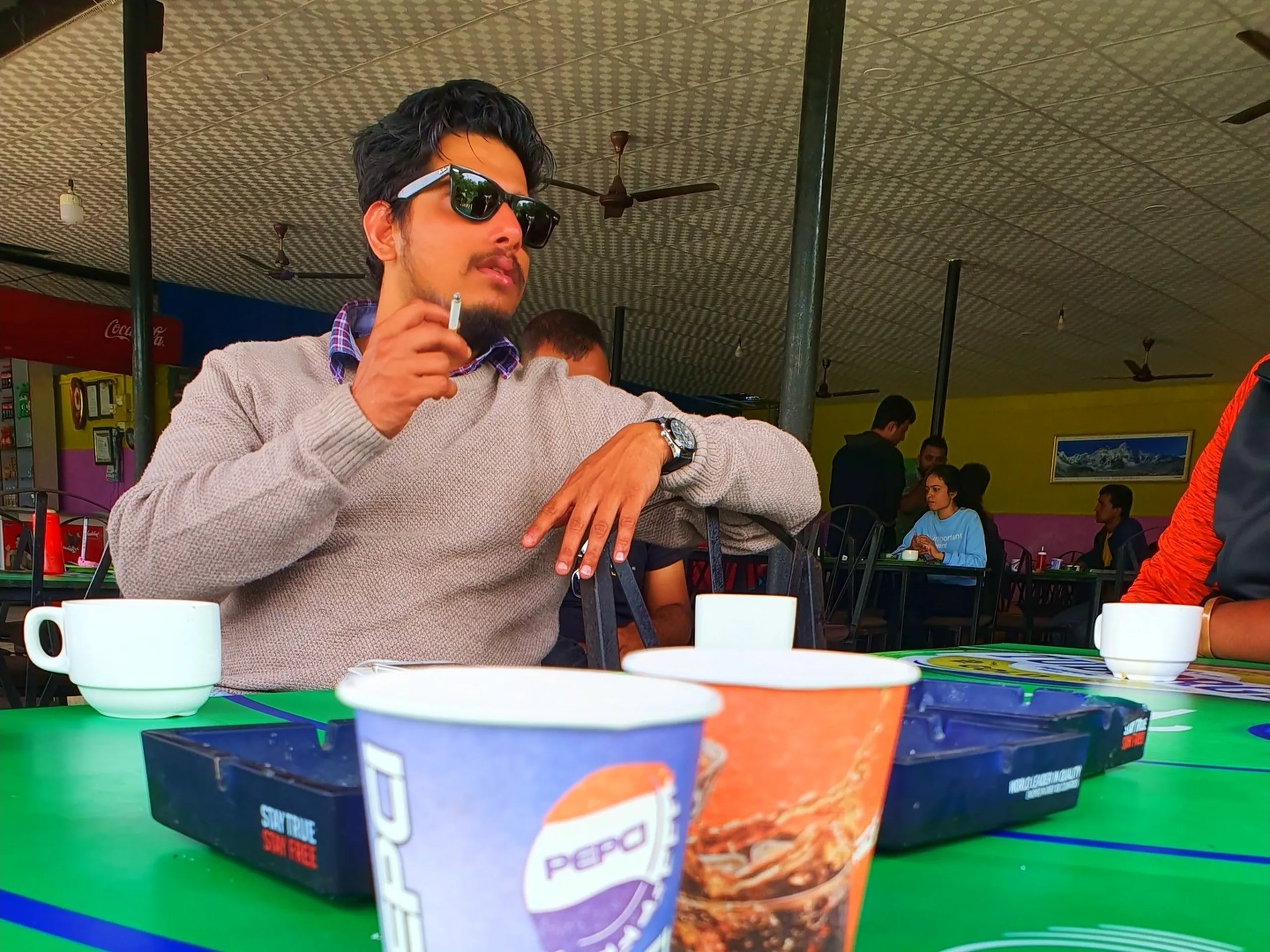 Young stylish male traveler in grey knit sweater black Ray-Ban wayfarer sunglasses wristwatch sitting at open-air roadside restaurant green table during travel stop Nepal white teacups Pepsi branded paper cups ashtray in foreground other diners visible in background yellow green walls Coca-Cola signage Nepal highway dhaba roadside restaurant Pokhara travel road trip pit stop blog