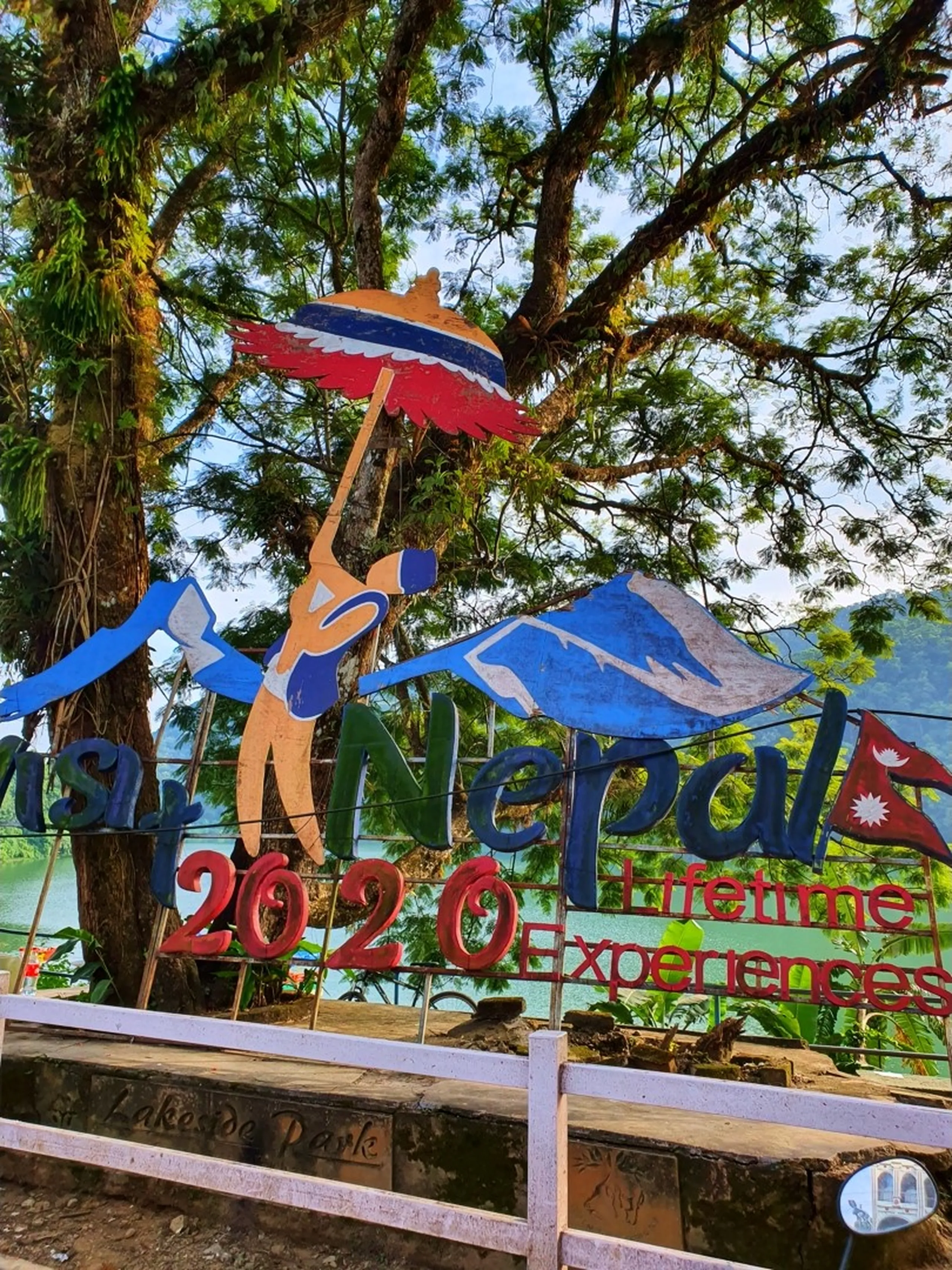 Colorful large outdoor promotional signage for Visit Nepal 2020 Lifetime Experiences tourism campaign located at Lakeside Park Pokhara Nepal featuring sky-blue stylized mountain climber figure holding flag with Nepal flag red white blue Himalayan peak silhouette turquoise Phewa Lake and lush green trees visible in background Pokhara Nepal tourism landmark travel photography visit Nepal travel blog