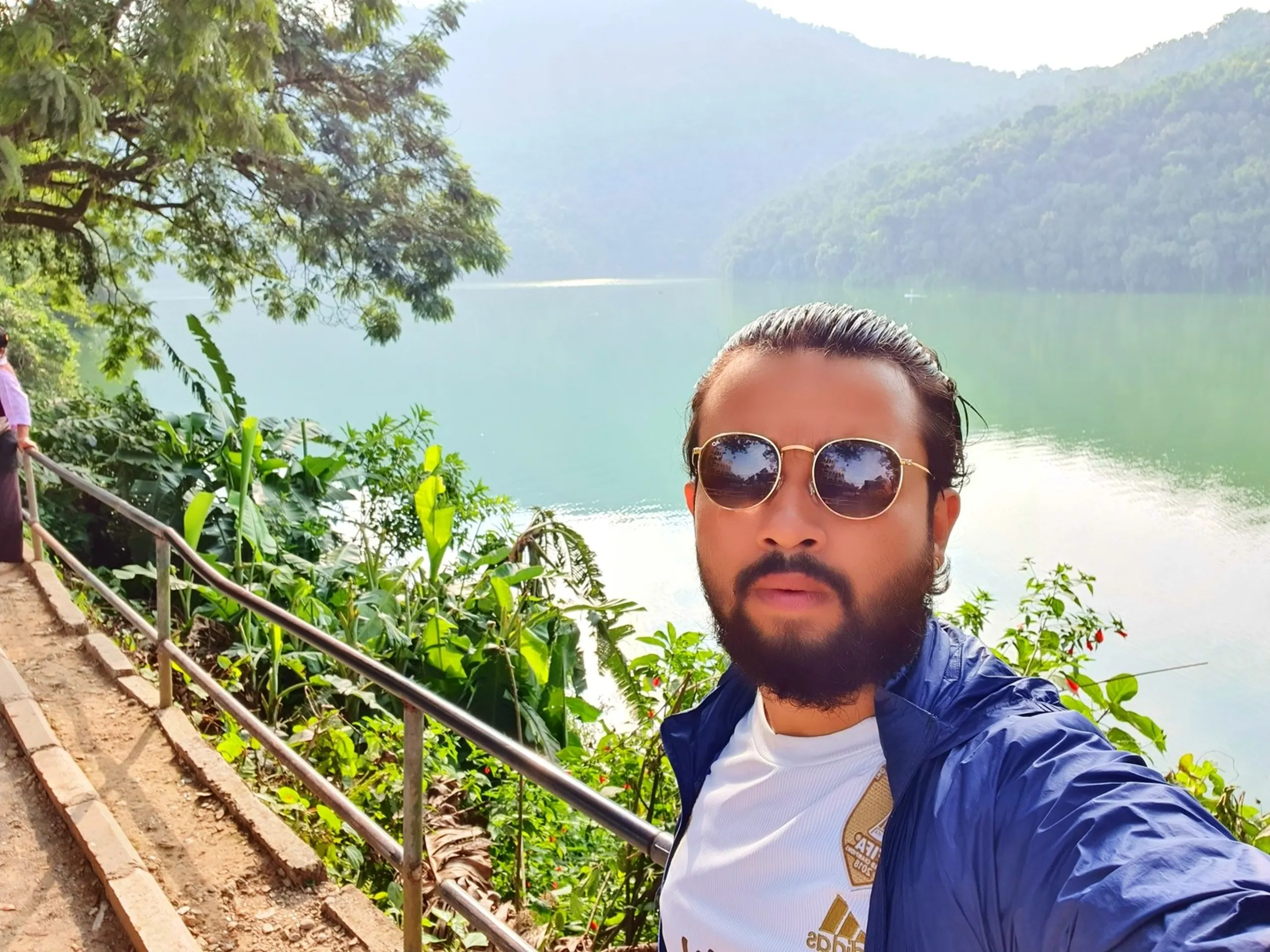 Close-up selfie of bearded male traveler with slicked-back hair gold-frame round mirror sunglasses blue waterproof jacket white Adidas t-shirt at Phewa Lake Pokhara Nepal lush tropical green banana leaves trees steel railing stone steps in foreground serene jade green lake water reflecting misty layered green Himalayan foothills in background another visitor partially visible Pokhara lakeside travel photography Nepal tourism must visit destination