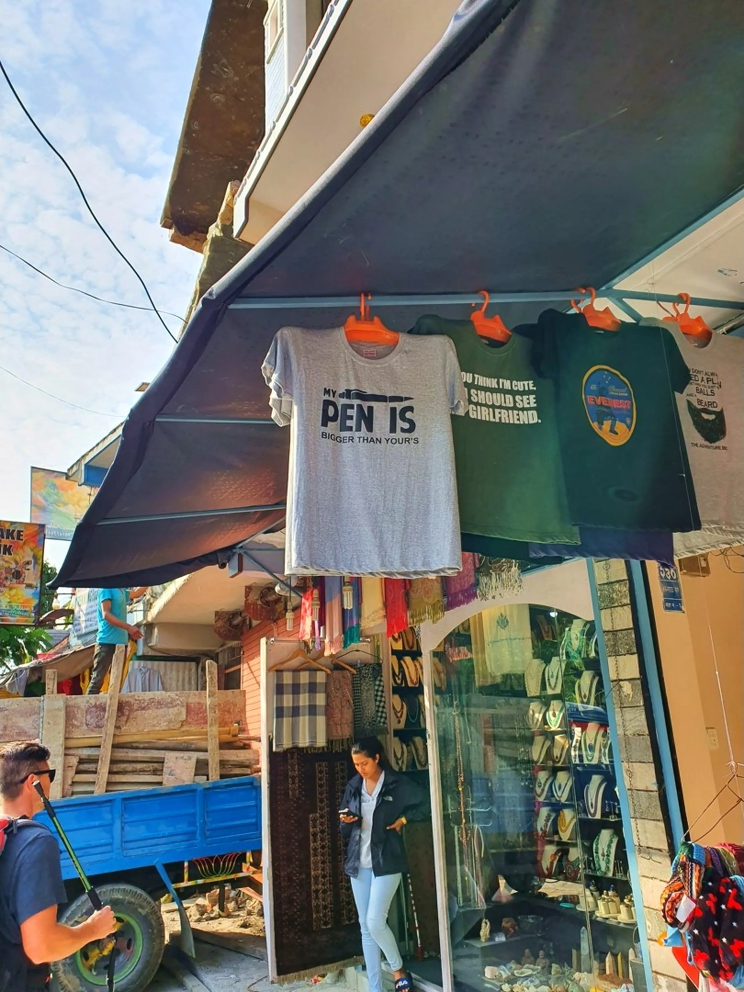 Funny novelty tourist t-shirt hanging outside Pokhara Lakeside souvenir shop Nepal reading My Pen Is Bigger Than Yours with pen graphic humorous travel merchandise alongside other novelty shirts and Everest logo tshirt green dark souvenir shop facade jewelry accessories scarves visible blue tractor parked below local woman in North Face jacket walking past Pokhara Lakeside market street shopping Nepal travel blog humor