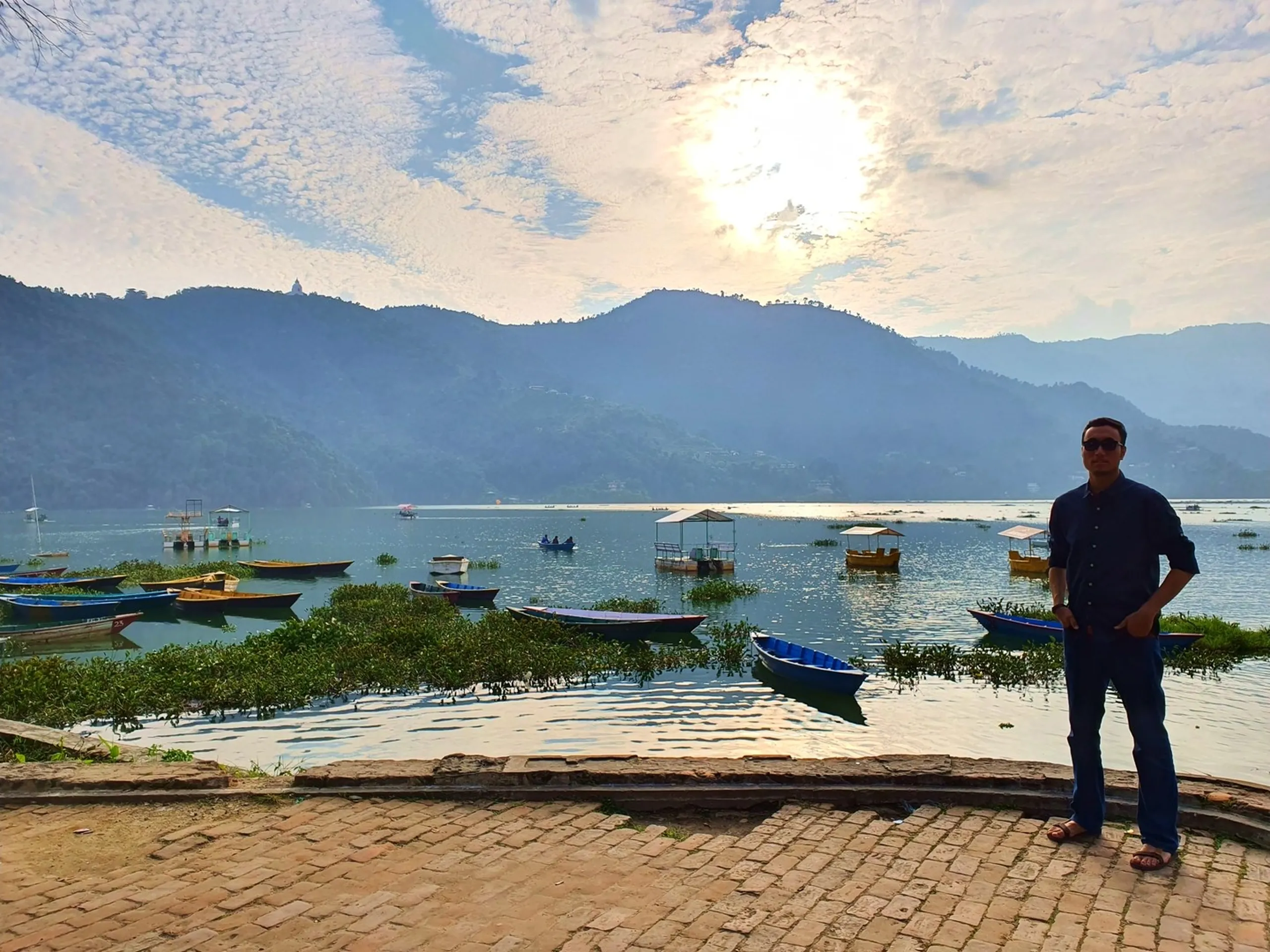 Male traveler in dark navy blue shirt jeans sandals sunglasses standing confidently at Phewa Lake Pokhara Nepal shoreline brick promenade with stunning panoramic view of multiple wooden rowing boats pedal boats moored on calm silver-blue lake water dramatic cloud formation sun behind clouds misty layered blue-green Himalayan foothills World Peace Pagoda hill visible Pokhara Nepal travel photography tourist landmark