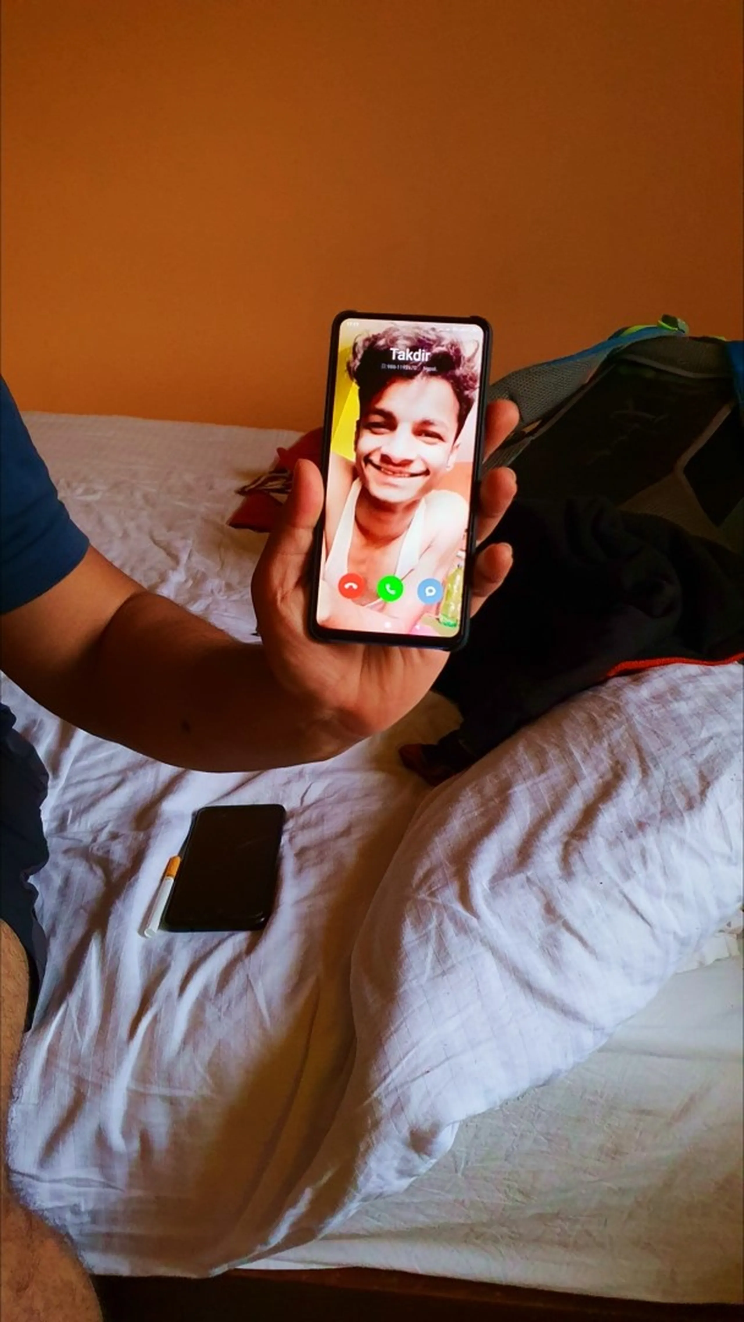 Close-up photograph of a smartphone held in hand displaying an incoming video call screen from a friend showing a young smiling boy's profile picture name visible on screen white bedsheets trekking backpack visible beside person on bed orange-painted hotel guesthouse room wall Nepal travel humor friends keeping in touch during trek adventure Annapurna Circuit Pokhara Nepal travel blog