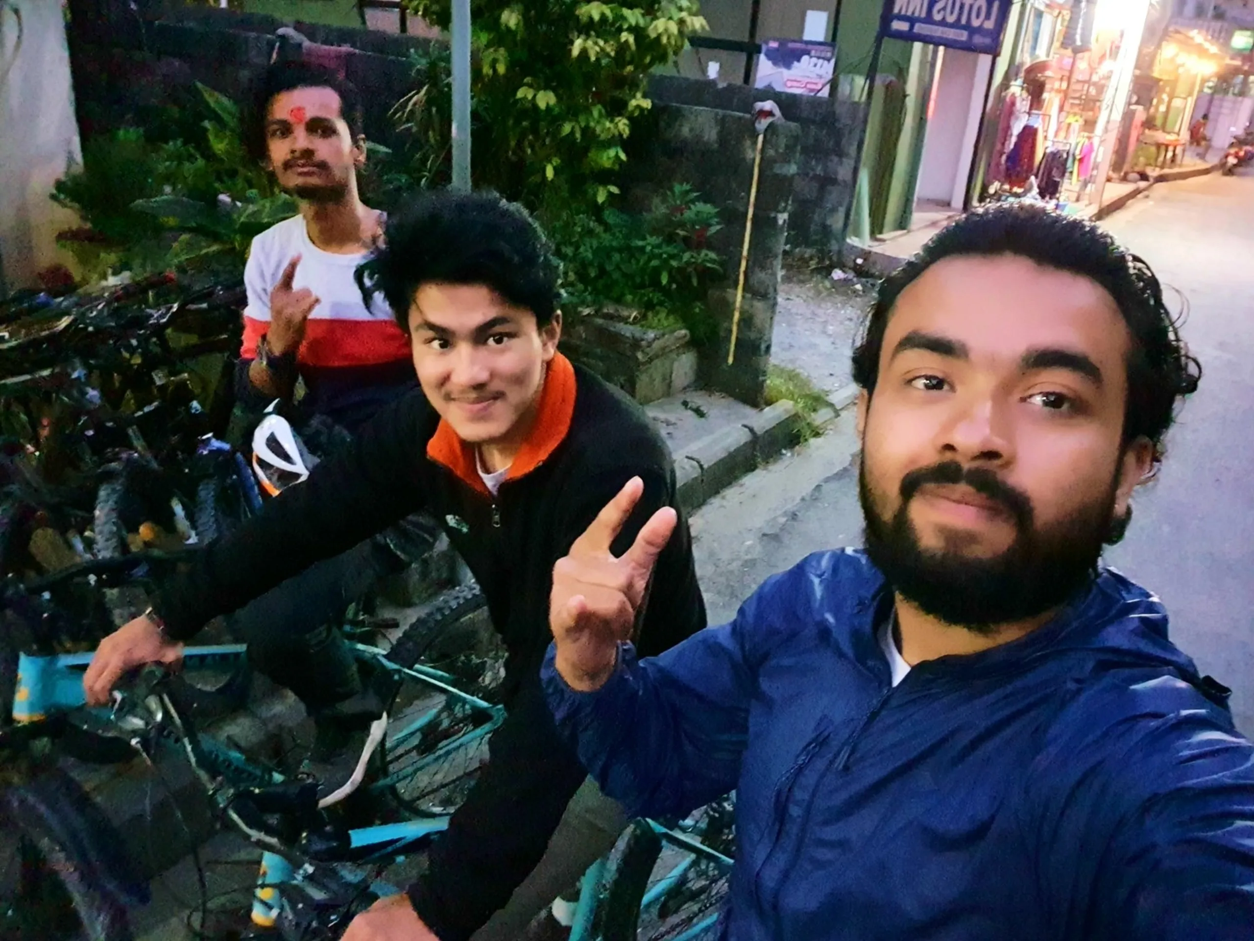 Night selfie of three young male friends cycling rented bicycles on dimly lit street in Pokhara Lakeside Nepal Lotus Inn signboard and lit shops visible in background one friend with red tikka mark on forehead white red striped t-shirt another in orange-collar black jacket giving peace sign bearded man in blue jacket in foreground Pokhara cycling nightlife local experience Nepal travel blog budget adventure