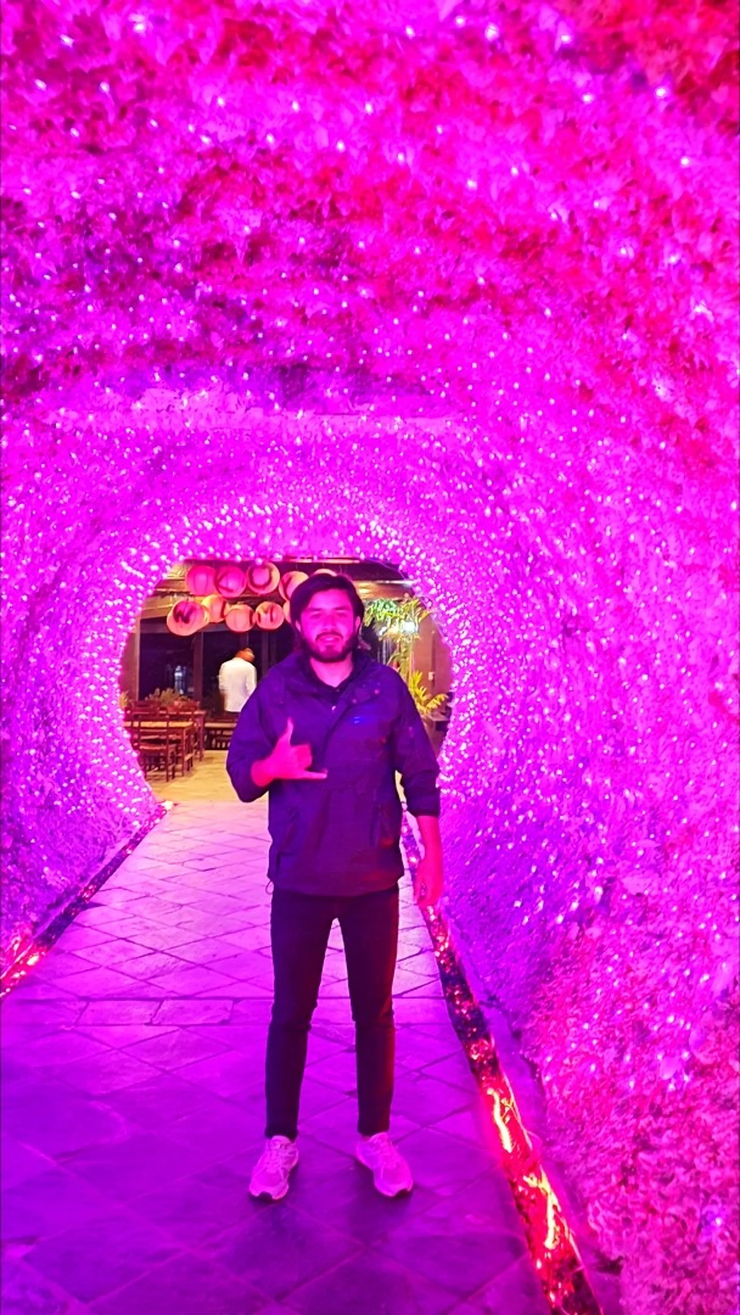 Smiling male traveler in dark navy jacket black cap white sneakers standing inside stunning illuminated pink magenta LED light tunnel archway at trendy Pokhara nightclub restaurant Nepal entire ceiling and walls covered in vivid hot-pink artificial flower lights creating dreamy glowing tunnel effect restaurant seating with warm lanterns visible through arch Pokhara nightlife Nepal travel blog clubbing Lakeside entertainment