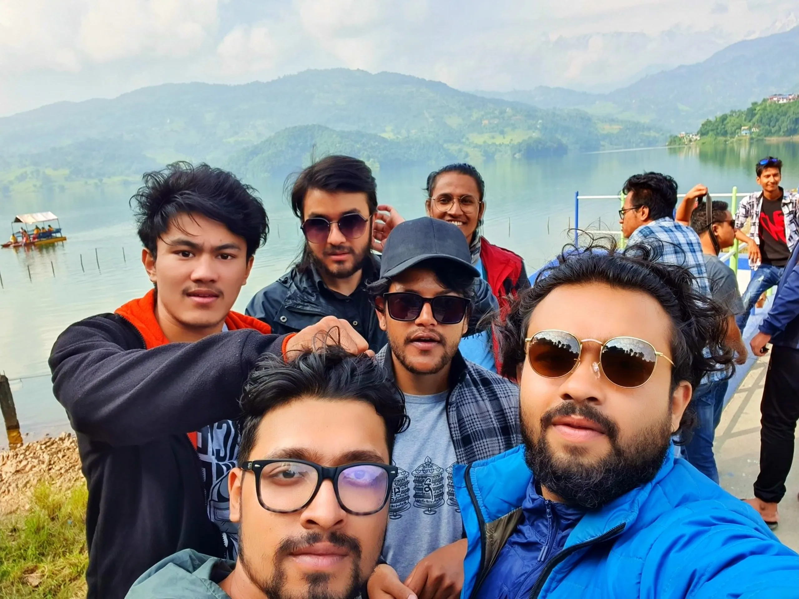 Final group selfie of six male friends at Begnas Lake Pokhara Nepal before returning to Kathmandu calm silver-blue lake water with moored covered boat raft and lush green rolling Himalayan hills visible in background overcast cloudy sky mixed casual travel outfits bearded man in blue jacket with round gold sunglasses man in checkered shirt dark sunglasses young man in orange collar black jacket Pokhara lakes Nepal travel blog farewell group photo