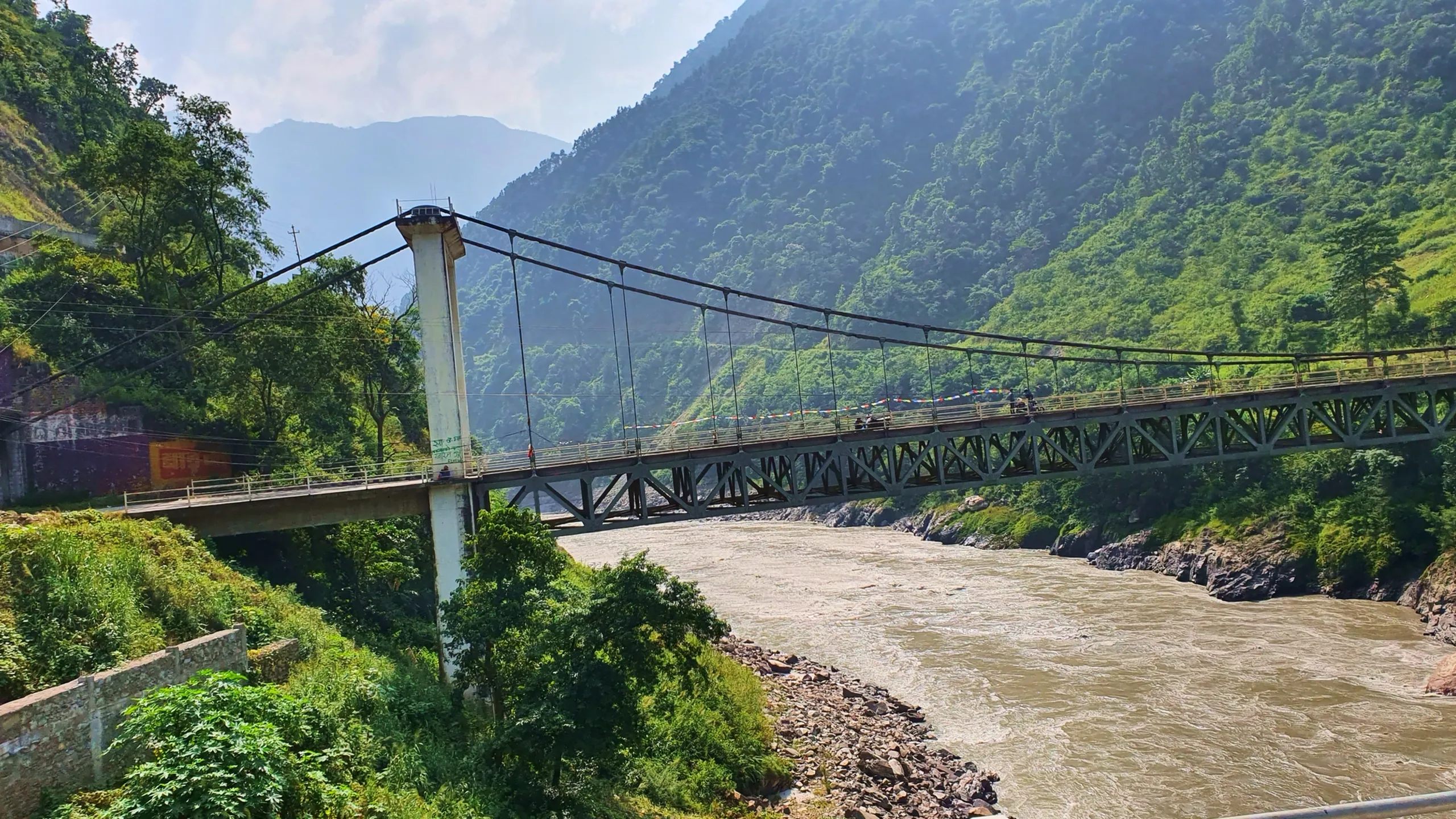 Iconic Mugling suspension bridge over powerful muddy brown Marsyangdi Trishuli River gorge Nepal dramatic wide-angle view of long steel truss bridge with tall white concrete pylon cable stays prayer flags strung along bridge railing dense lush green tropical jungle forest covering steep Himalayan foothills on both sides river rocky shores in foreground Mugling Nepal famous road trip landmark Kathmandu Pokhara highway bridge travel photography
