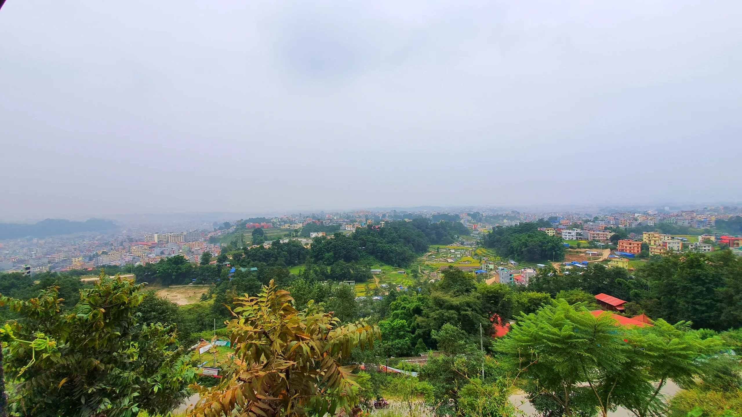 Wide elevated panoramic view of Kathmandu valley Nepal from hilltop showing sprawling dense urban city spreading across green valley colorful multi-storey buildings orange red yellow blue rooftops lush green trees terraced green paddy fields in foreground overcast hazy grey monsoon sky Nepal capital city return from Annapurna Circuit trek Kathmandu cityscape travel photography back to civilization Nepal adventure end